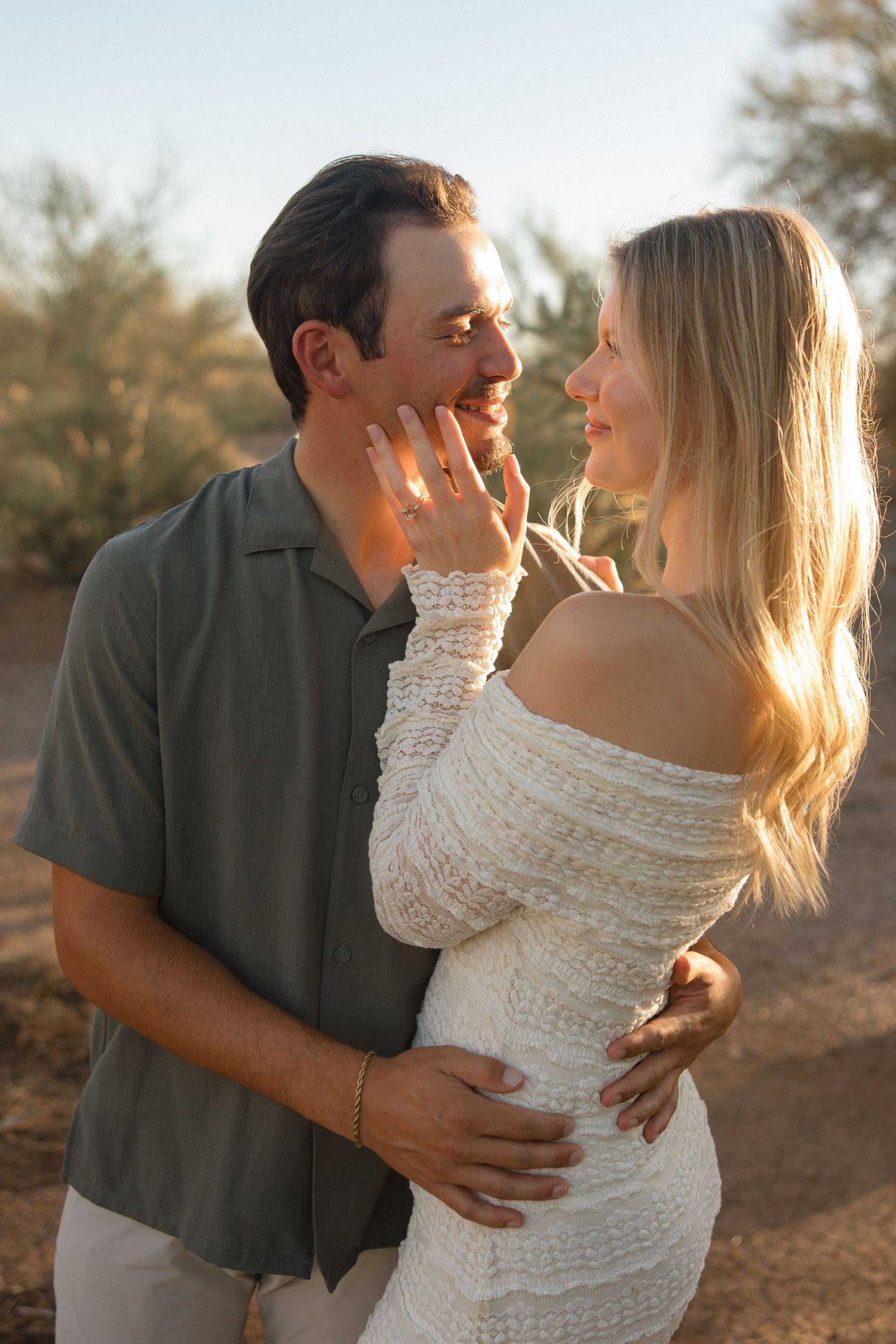 A couple embraces at golden hour, the woman in a white lace dress touching the mans face, showing an engagement ring.