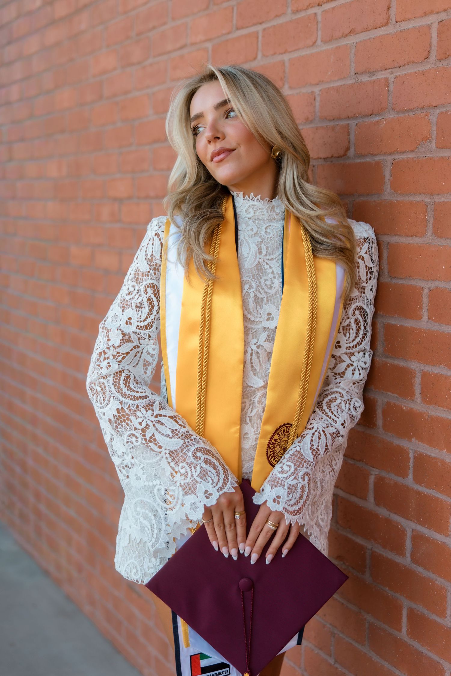 Blonde woman in white lace dress with gold graduation stole holds maroon cap against brick wall.