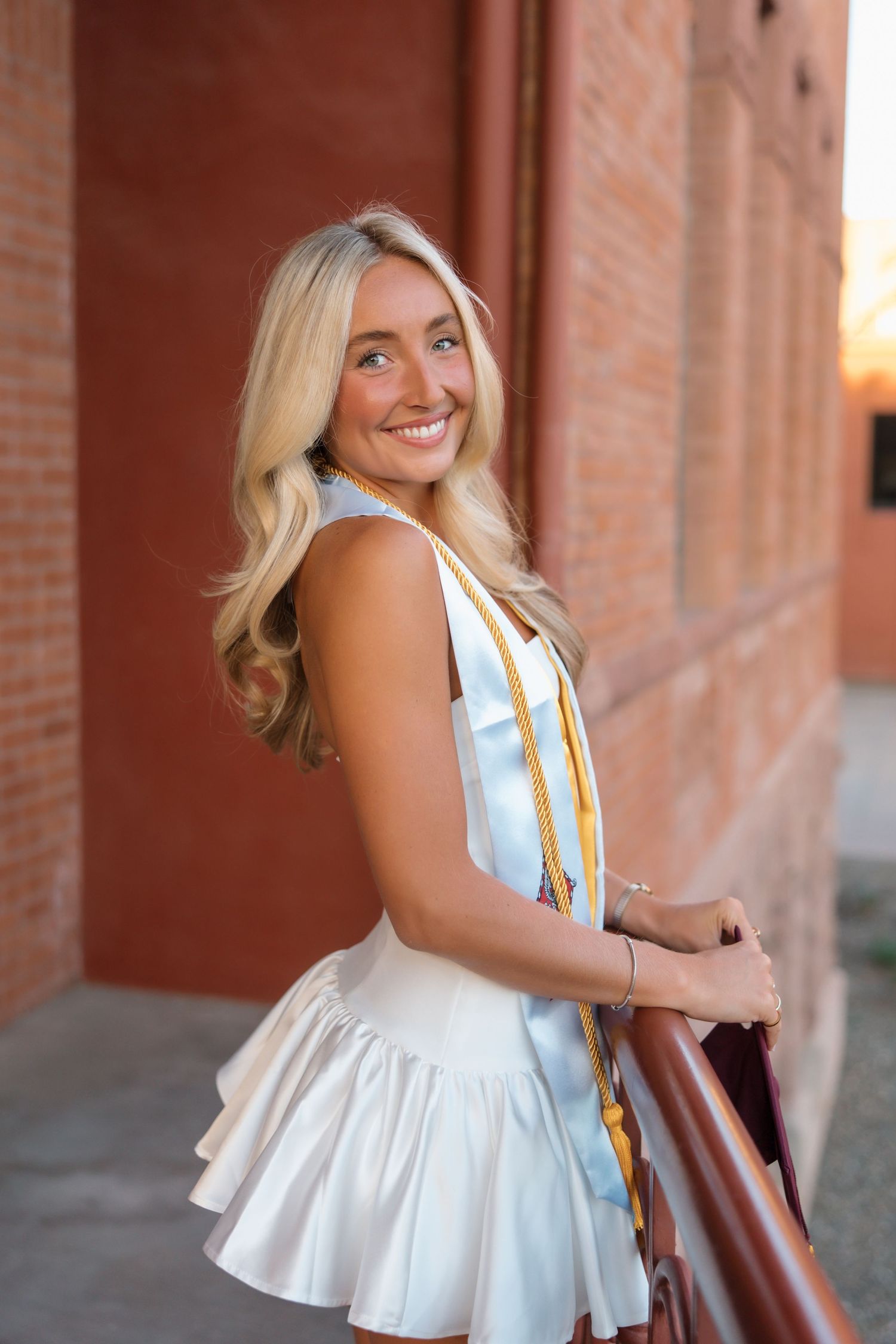 Smiling blonde woman in white ruffled dress with graduation stoles leaning on railing at sunset.