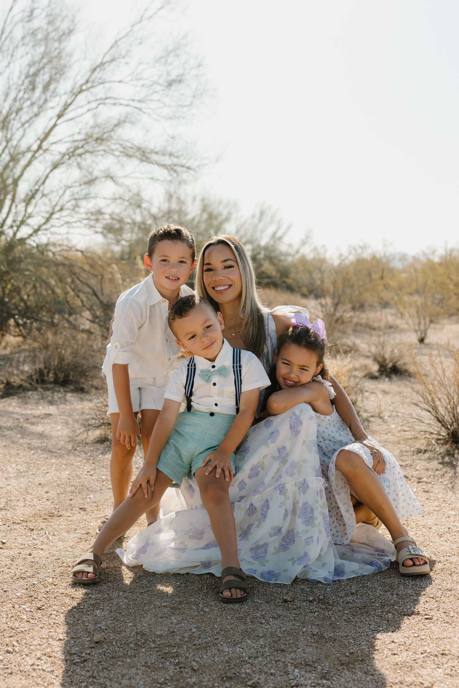A woman sits outdoors in a desert landscape with three children posing together for a family portrait.