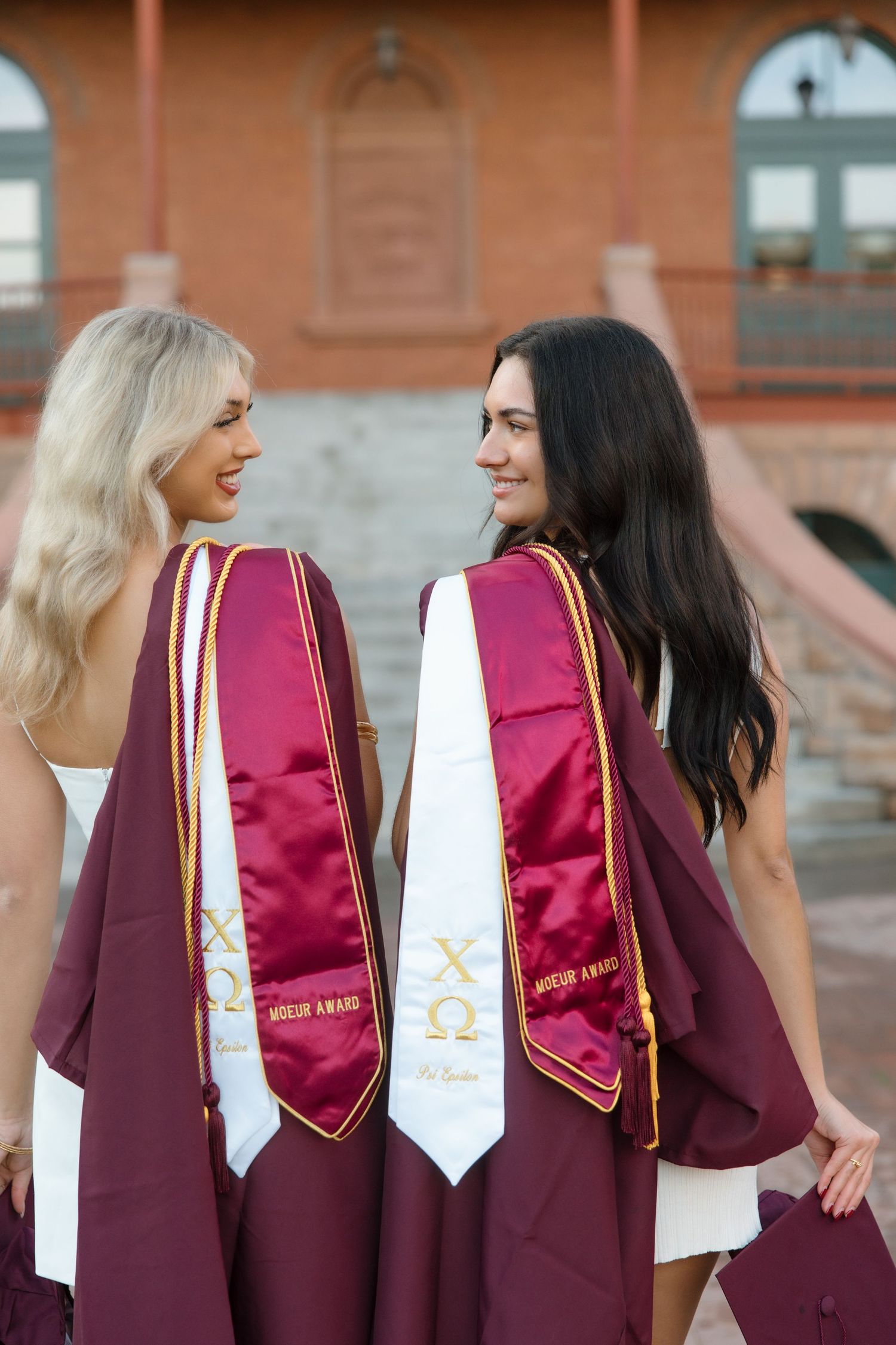 Two smiling ASU Chi Omega graduates in maroon robes and stoles facing each other outside on campus.