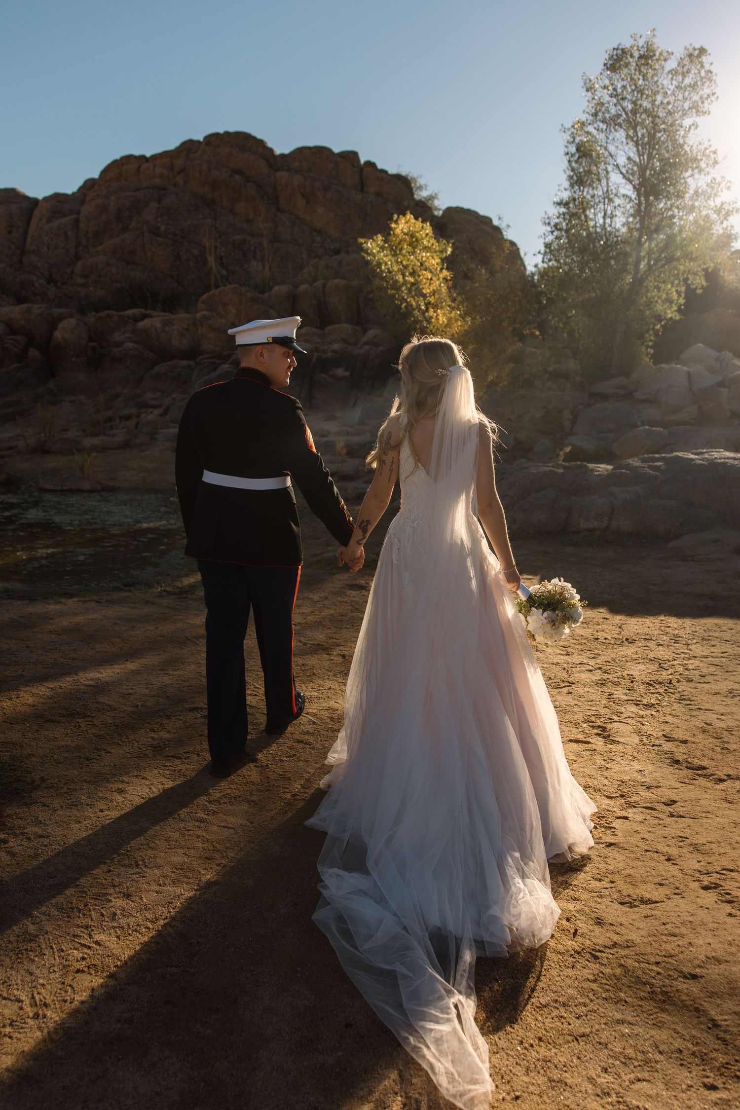 Marine groom in dress uniform holds hands with bride in flowing white gown and veil at scenic outdoor wedding.