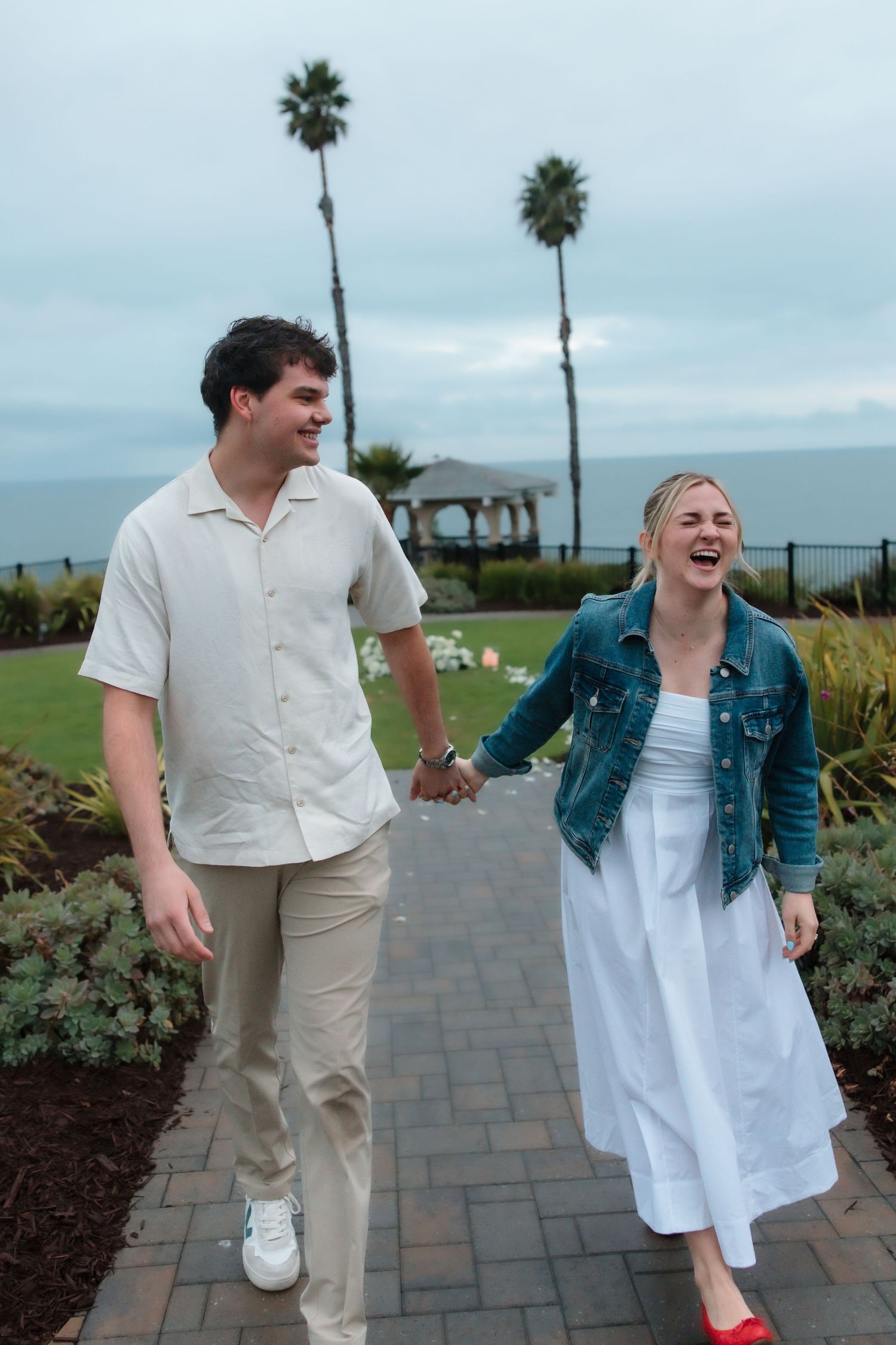 Joyful engaged couple holding hands walking on garden path with palm trees and ocean view.