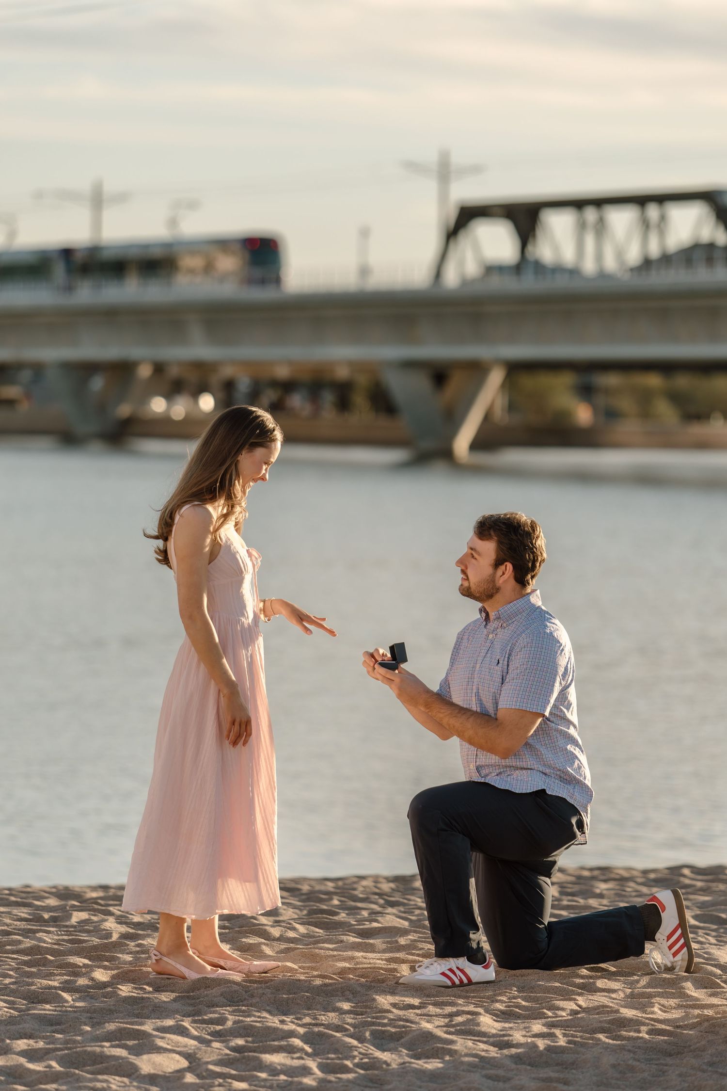 Man proposing on one knee to woman in pink dress on sandy beach at sunset with bridge in background.