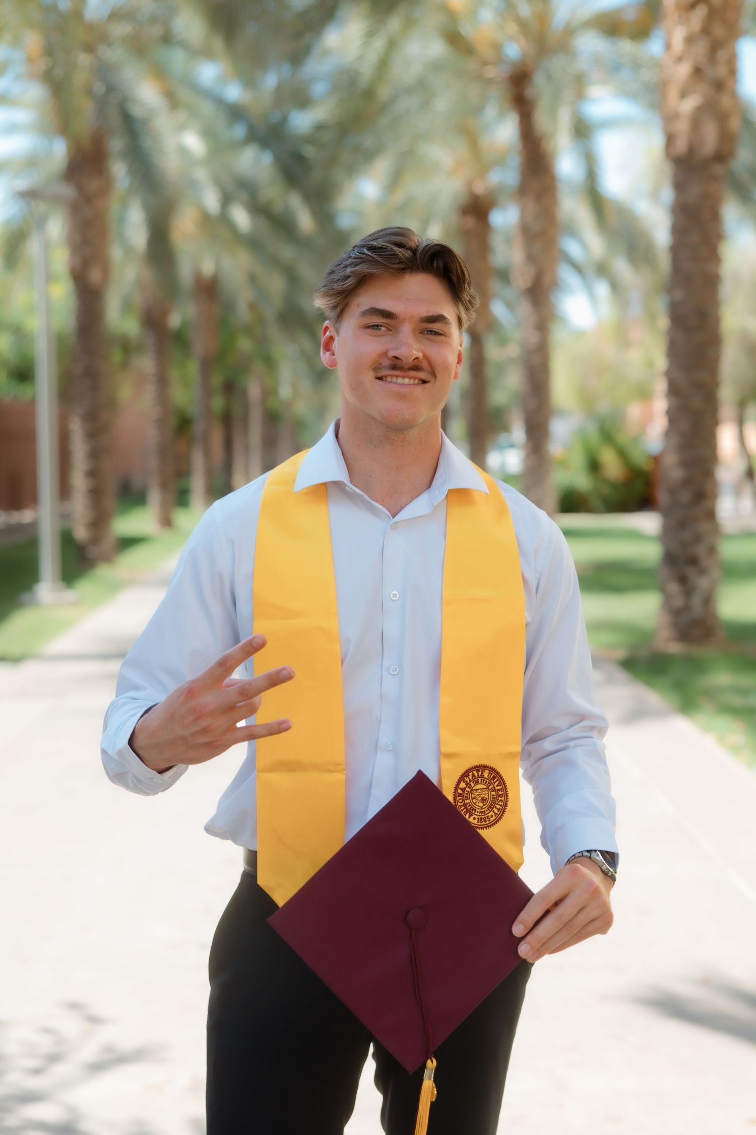 Young male ASU graduate smiling with gold stole and maroon cap among palm trees on campus.