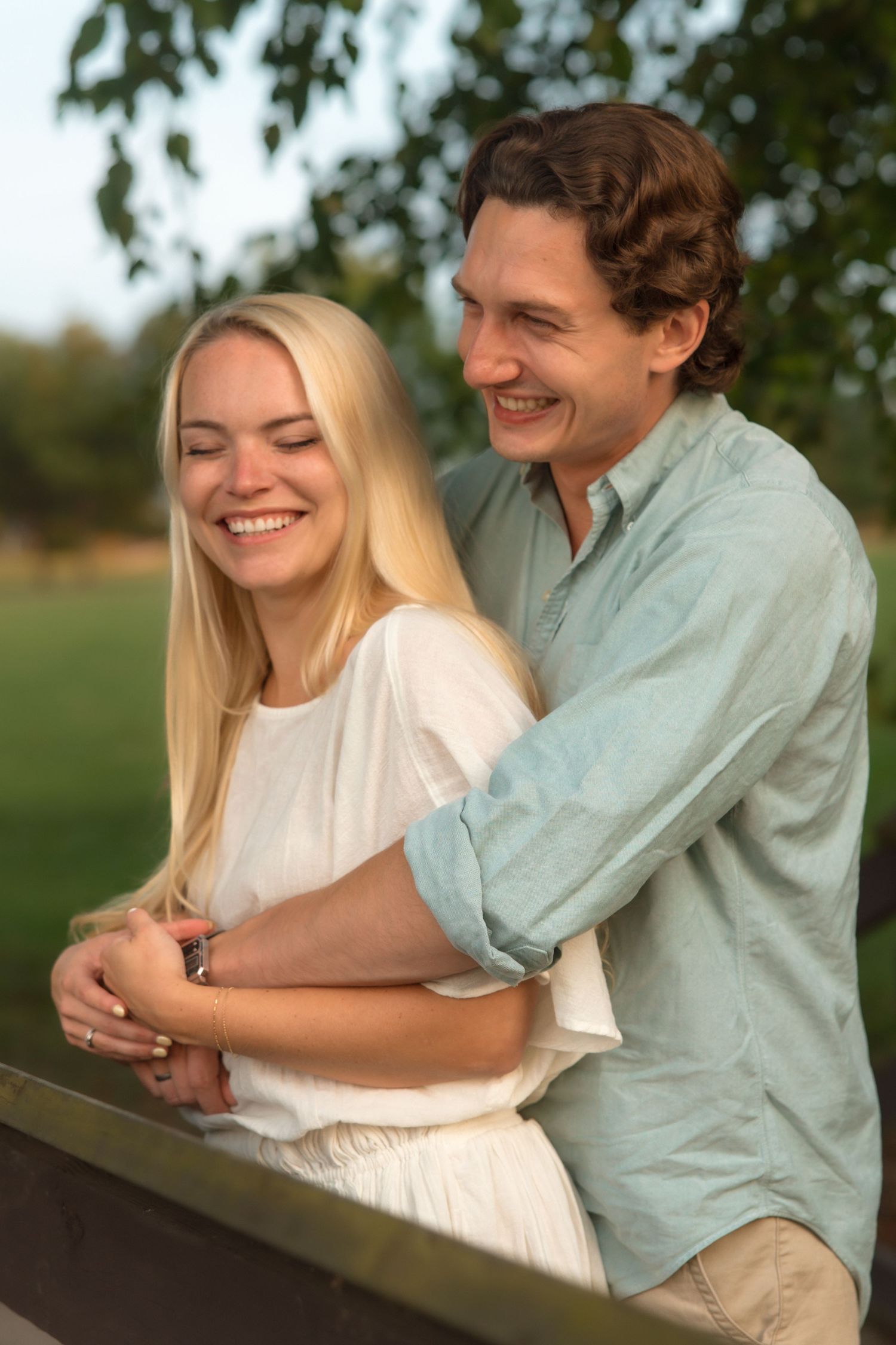 A joyful couple laughs together on a wooden bridge, the man embracing the woman from behind.