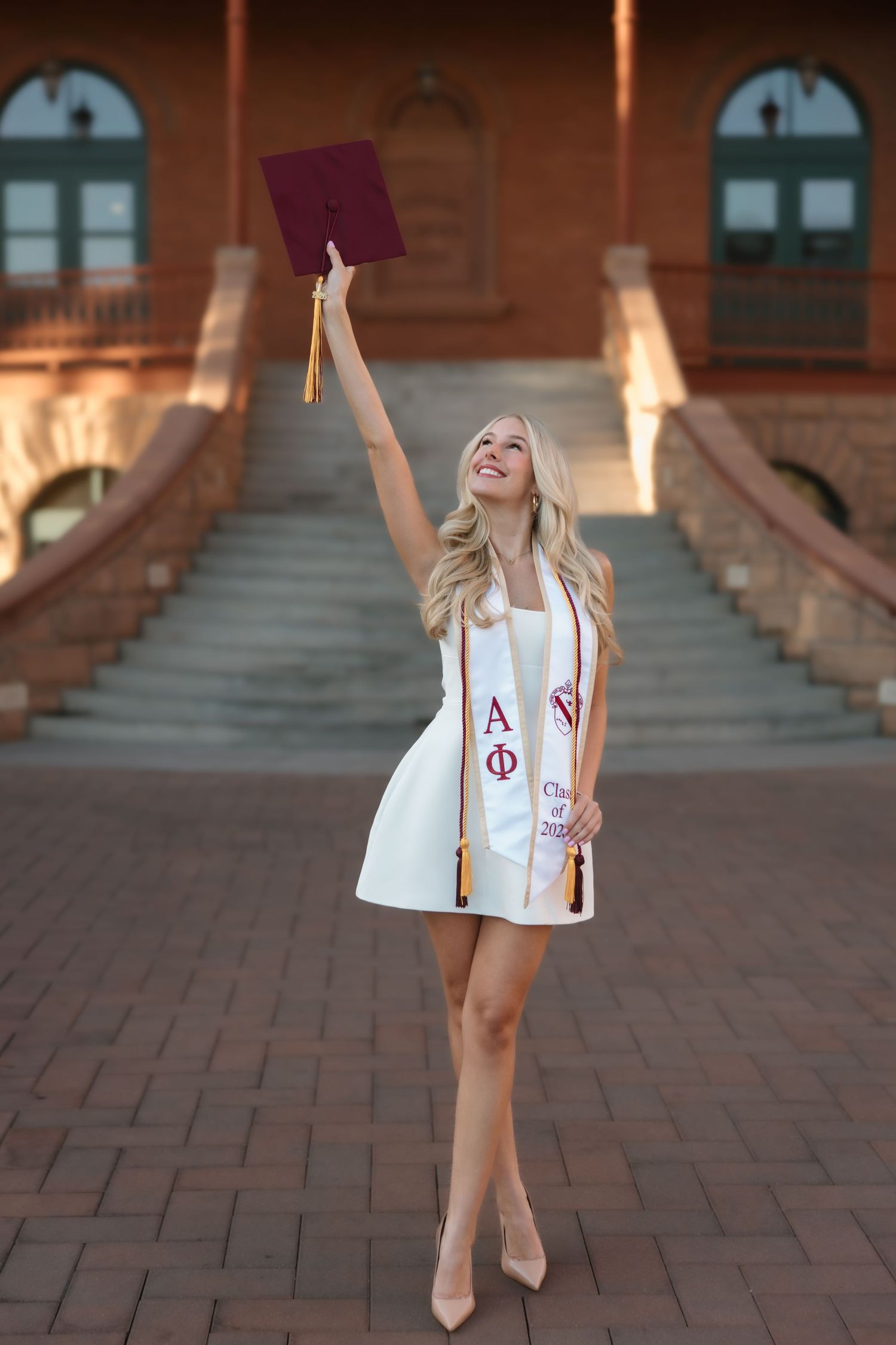Joyful female graduate raises maroon cap wearing white dress with Alpha Phi stole outside ASU brick building.