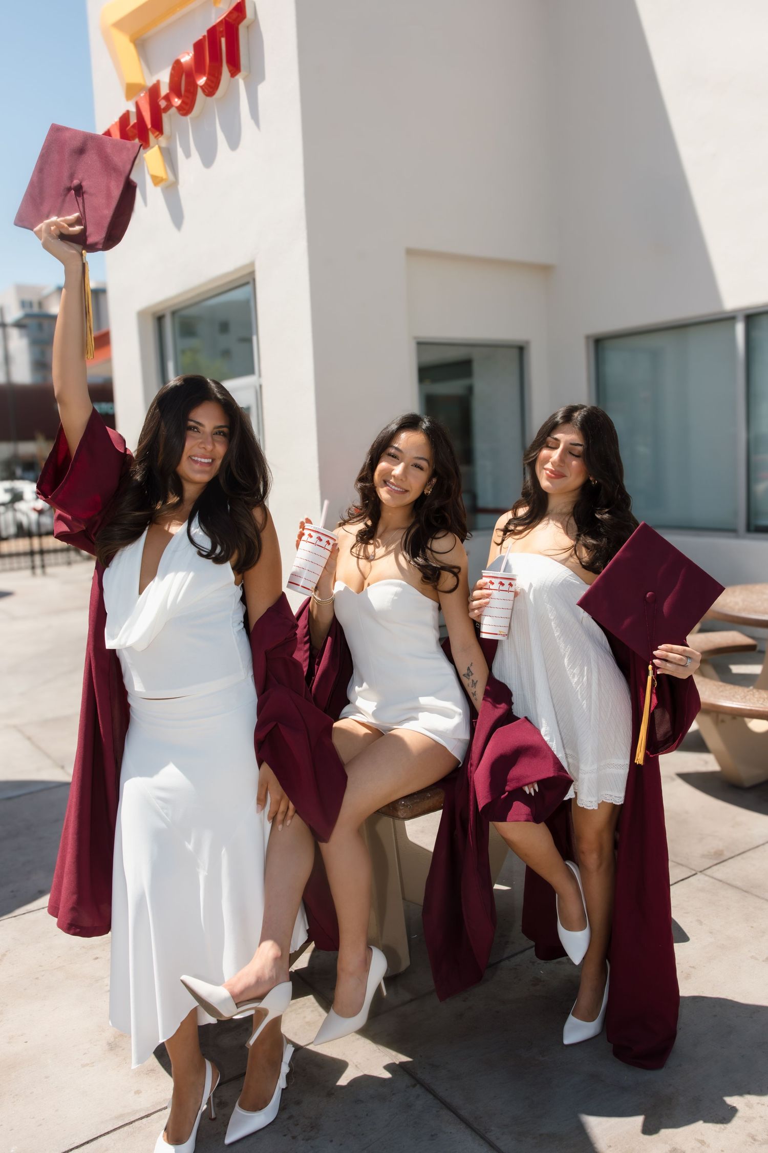 Three female graduates in burgundy robes sit outside In-N-Out Burger holding caps and drinks in the sun.