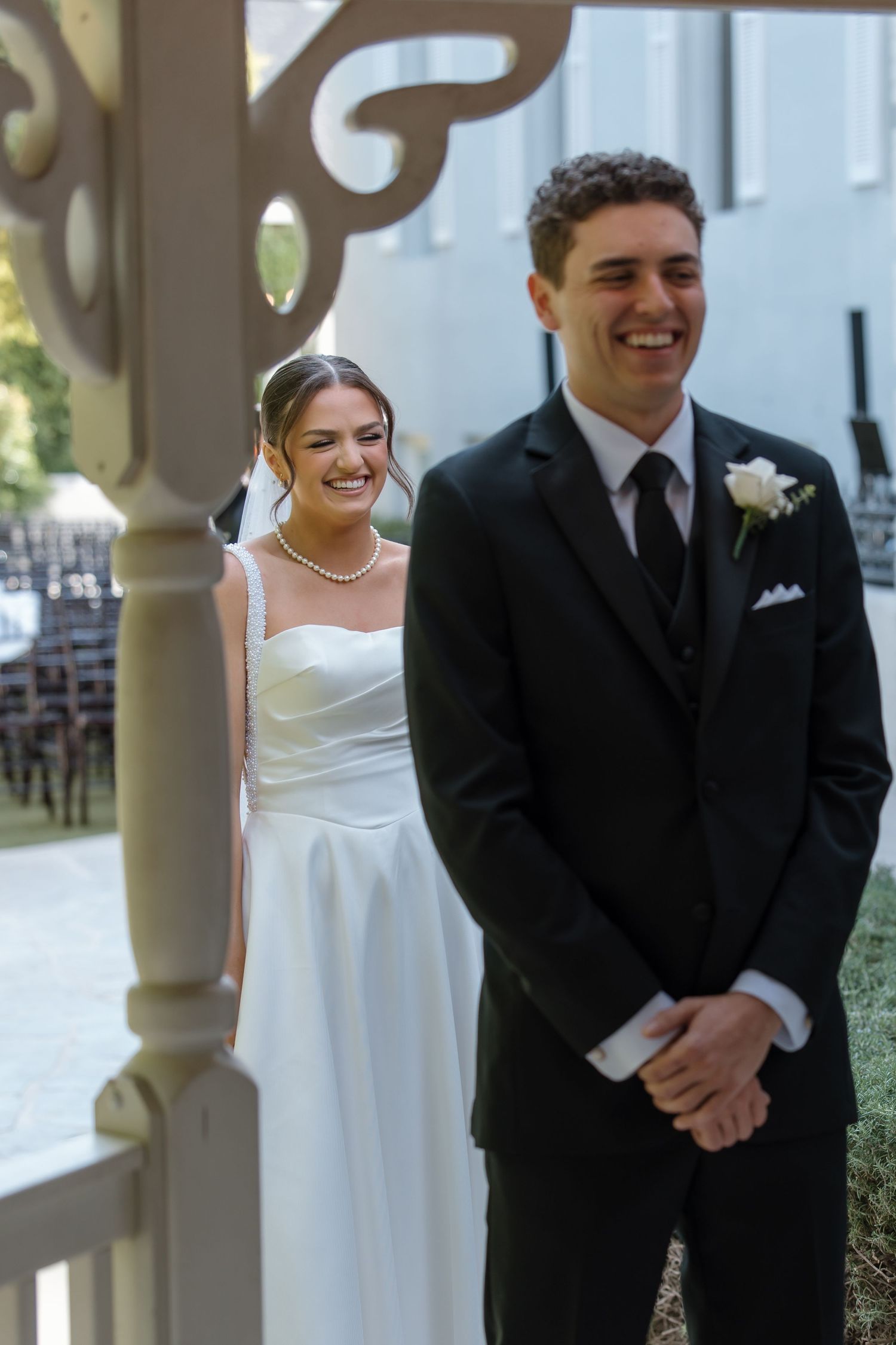 Smiling bride in white gown and pearl necklace standing behind groom in black suit during first look.