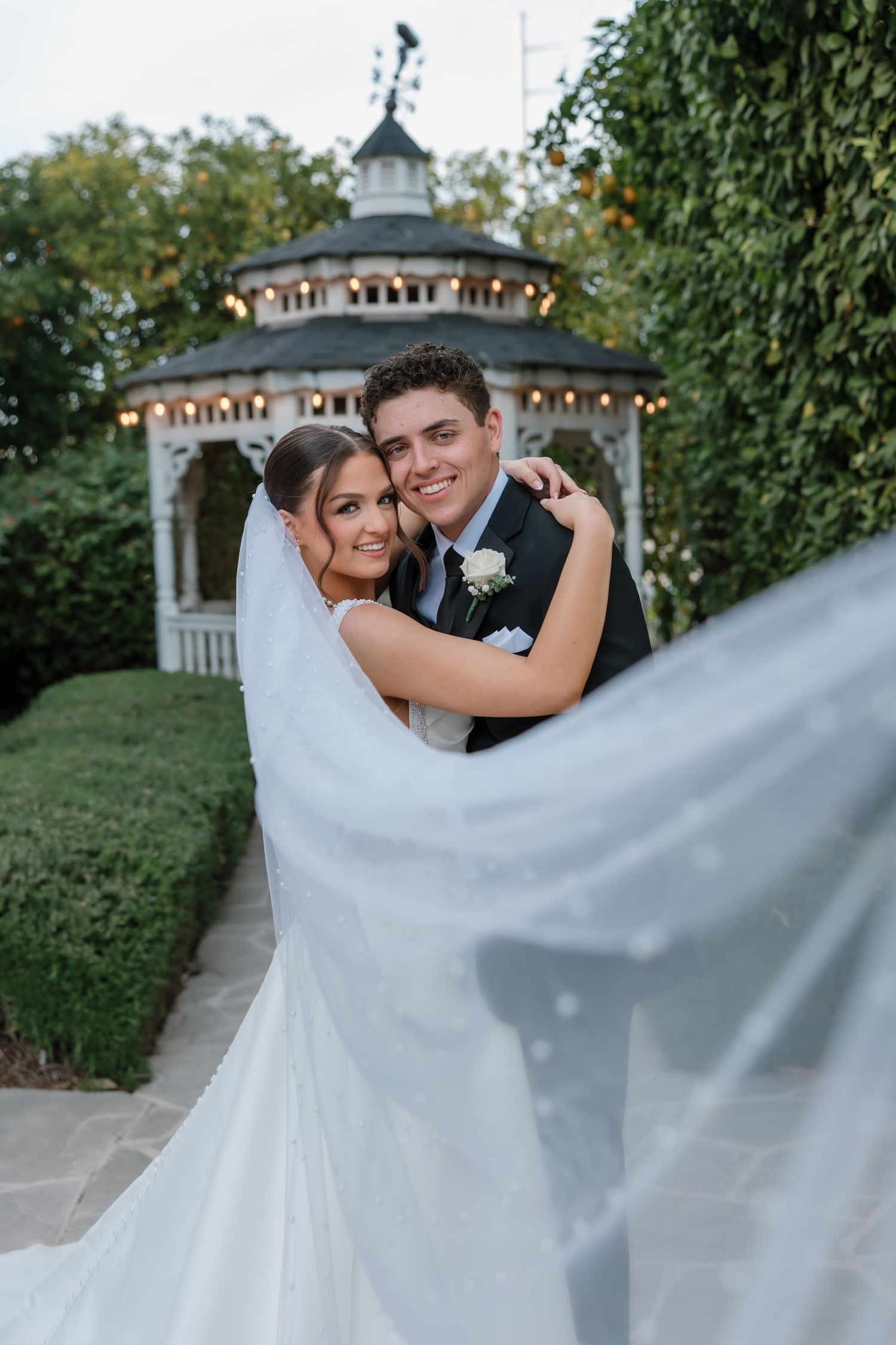 Bride and groom embrace in front of a lit gazebo as her white veil flows in the breeze.