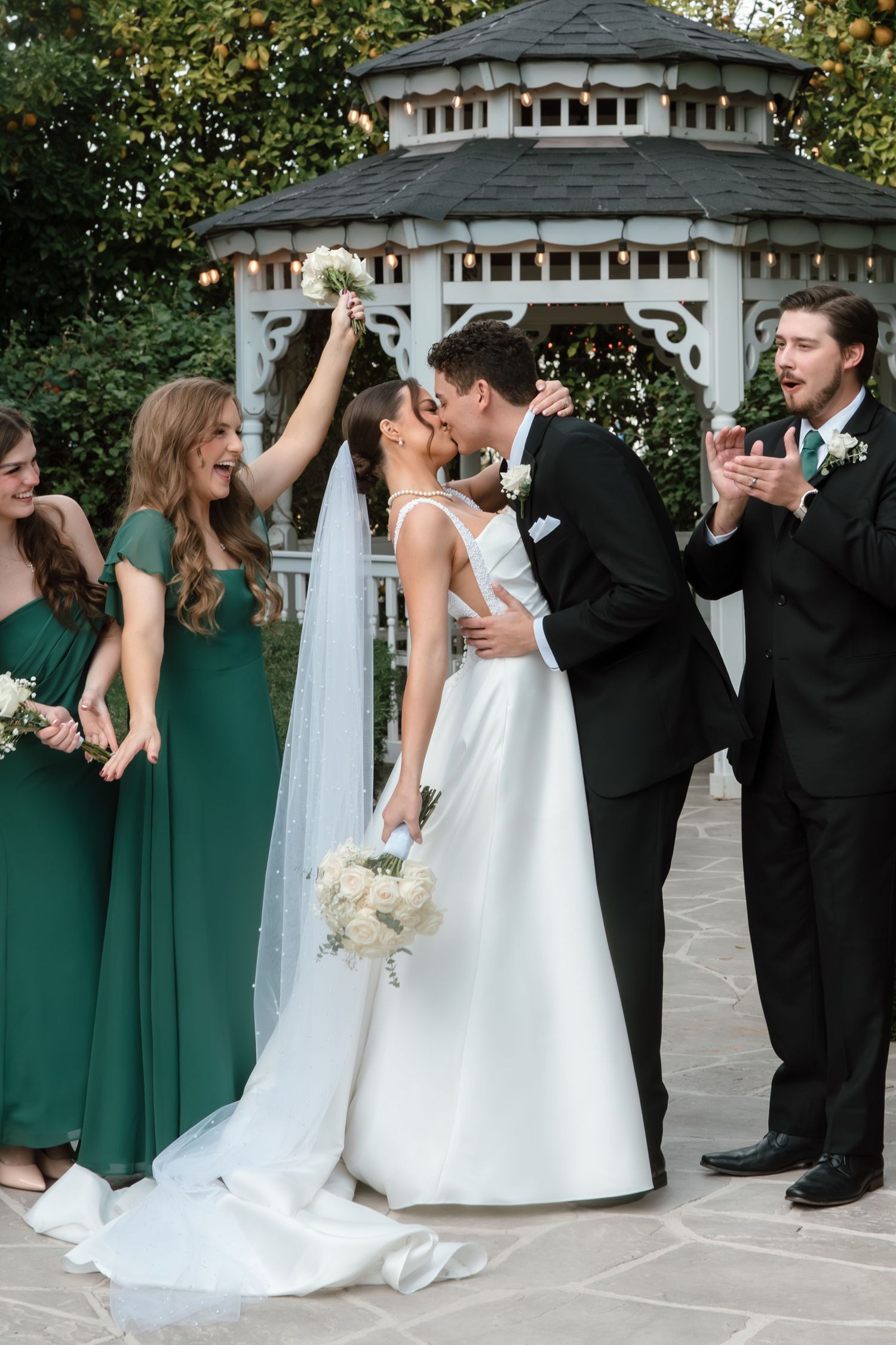 Bride and groom kiss as bridesmaids in emerald green celebrate with bouquet raised at gazebo wedding.