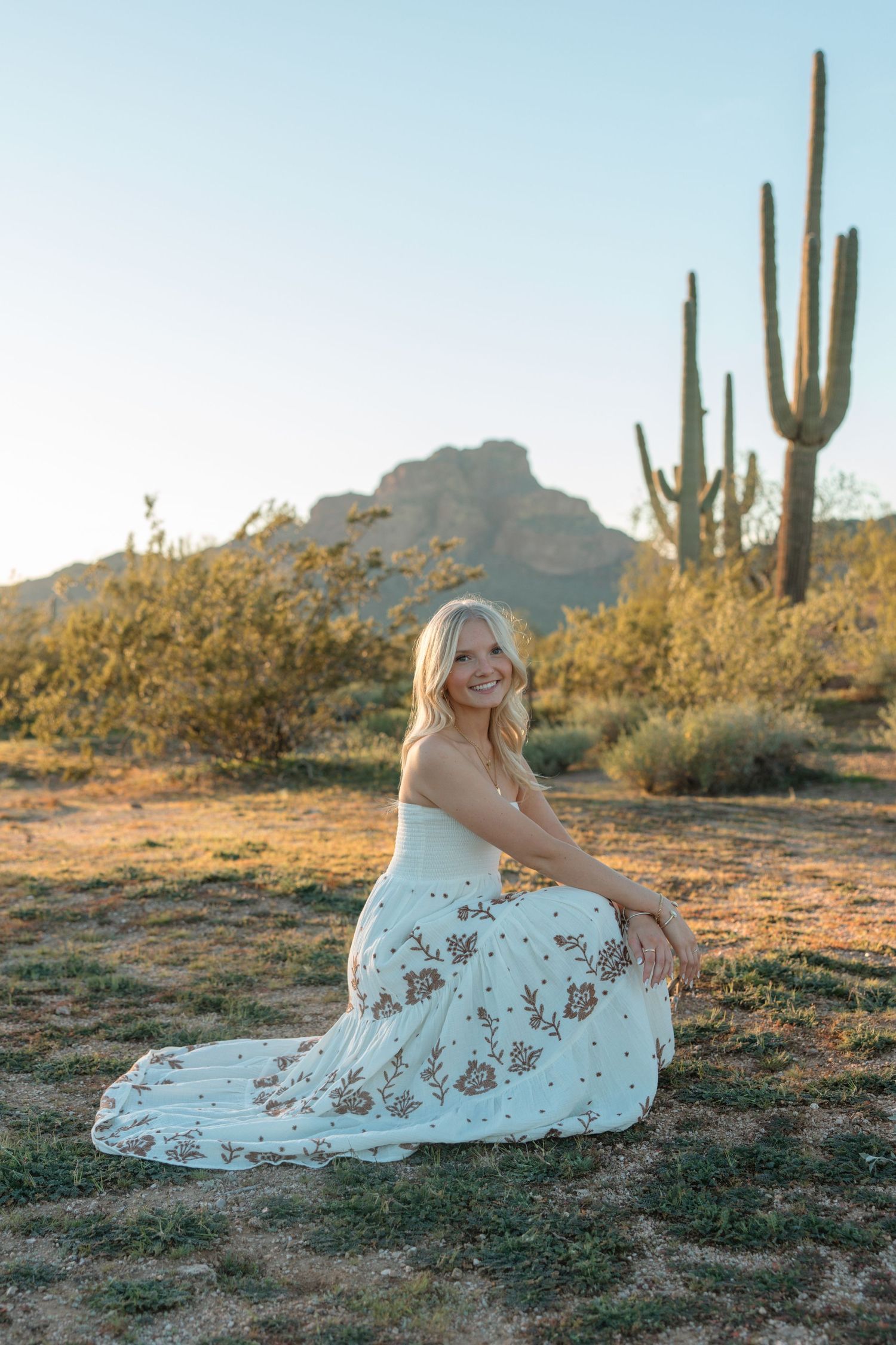 Blonde woman in white floral dress sitting gracefully in desert with saguaro cactus backdrop.