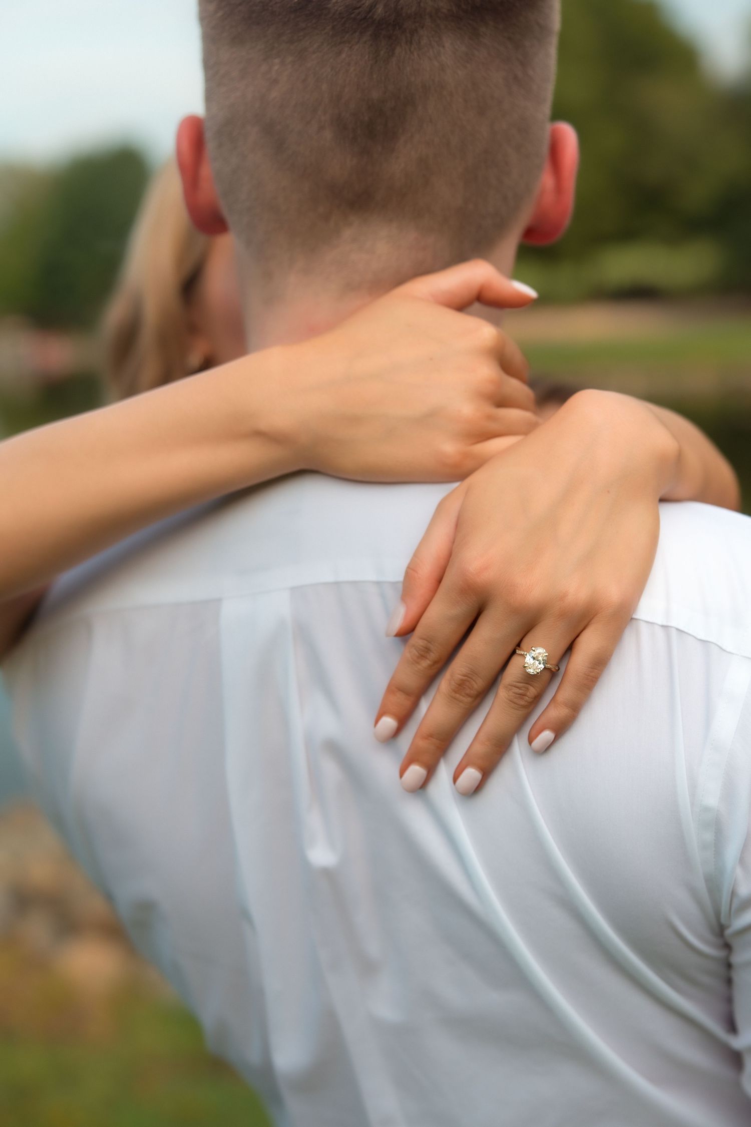 Woman with engagement ring embracing man from behind, showcasing gold diamond ring on her finger outdoors.