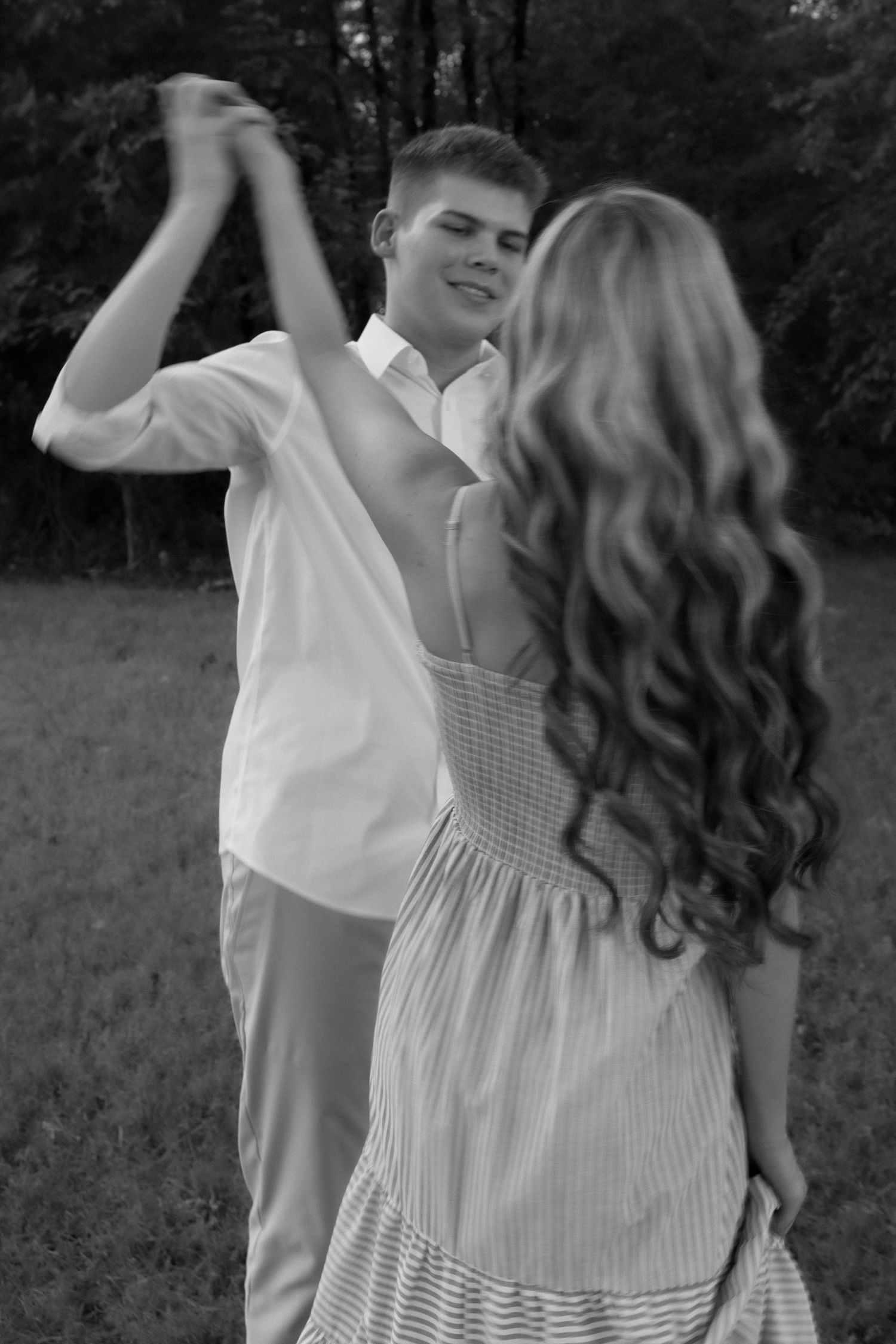 Black and white photo of a young couple dancing outdoors, man in white shirt smiling while twirling woman in striped dress.