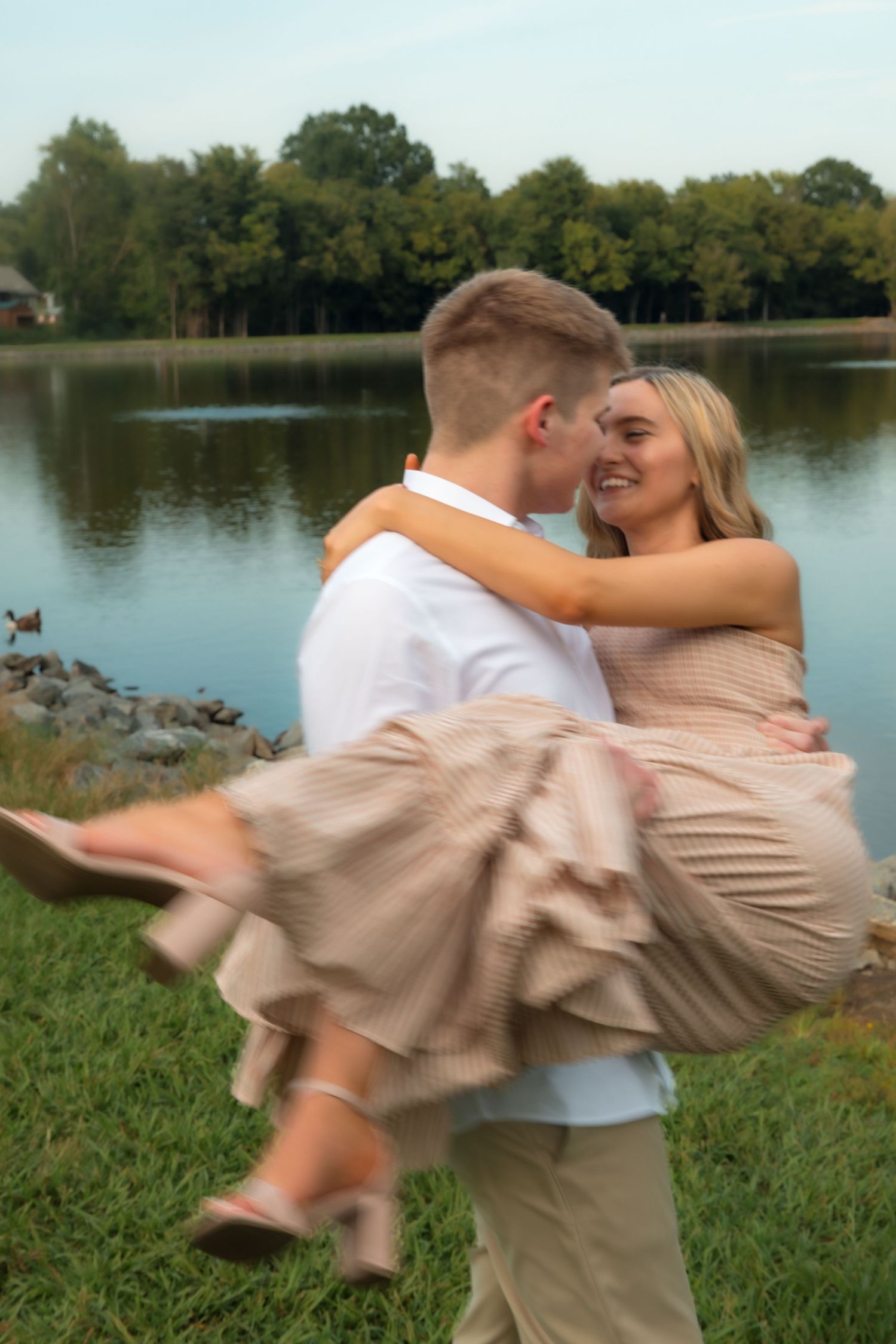 Young couple sharing a romantic moment by a lake, man lifting woman in a beige dress outdoors.