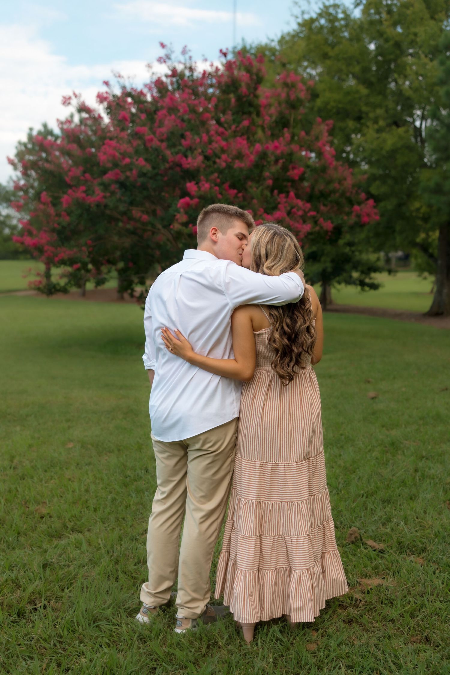 Couple sharing a romantic kiss in a lush green park with blooming pink crepe myrtle trees in the background.