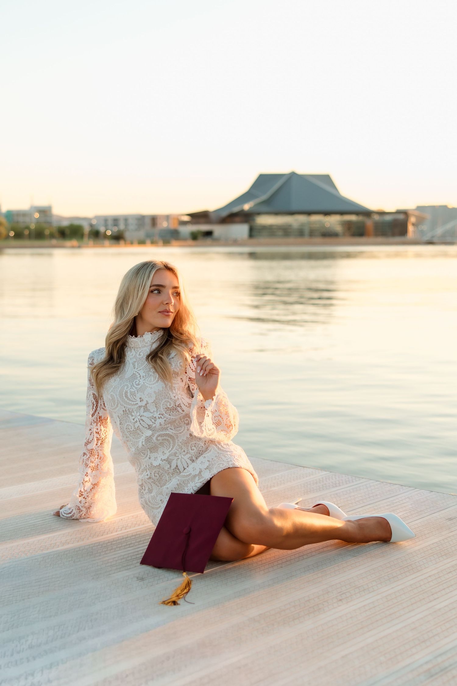 Blonde woman in white lace dress sits on dock at golden hour with maroon graduation cap.