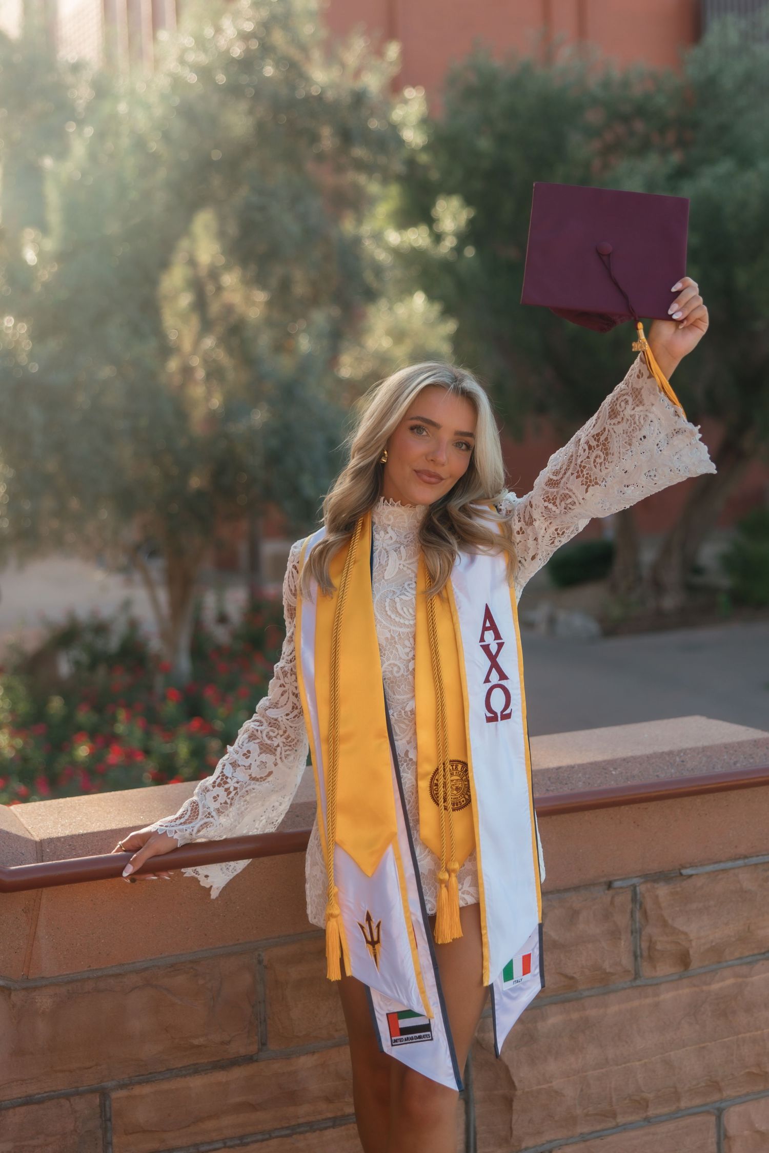 Smiling graduate holds maroon cap wearing Alpha Chi Omega stole and gold honor sash on campus.