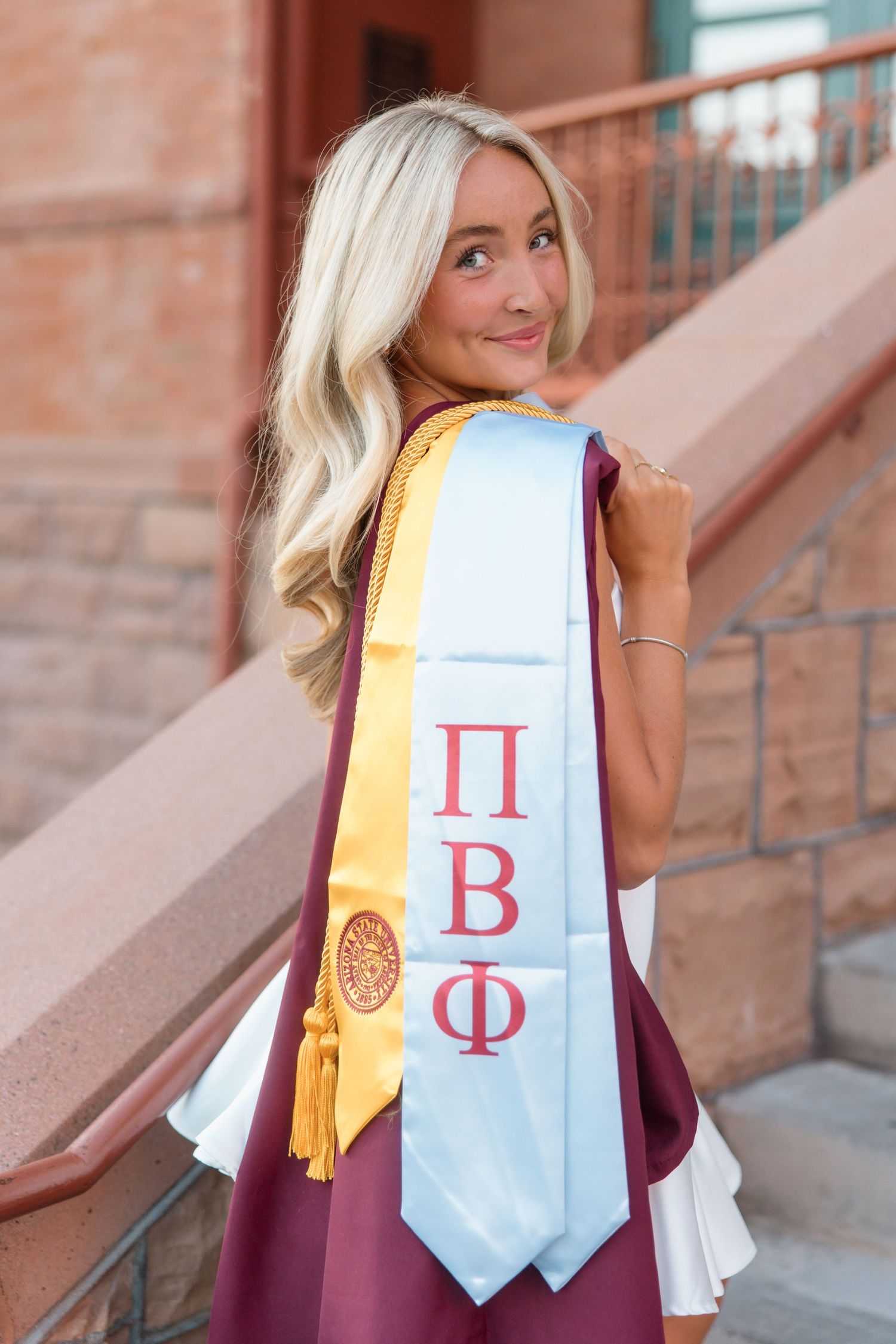 Blonde graduate showing Pi Beta Phi sorority stole and gold tassel on maroon graduation gown.