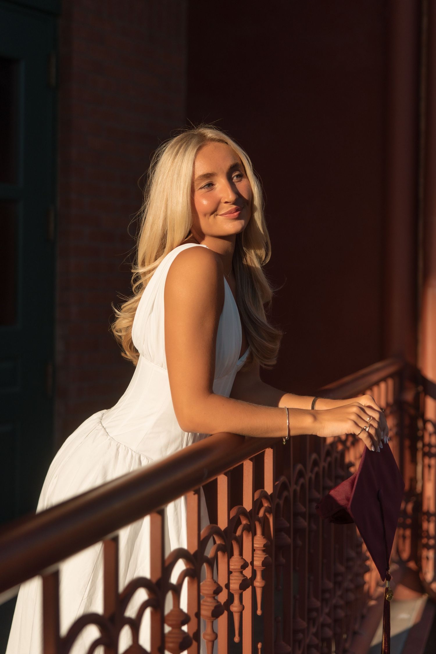Blonde female graduate in white dress leans on ornate railing in warm golden sunset light, smiling softly.