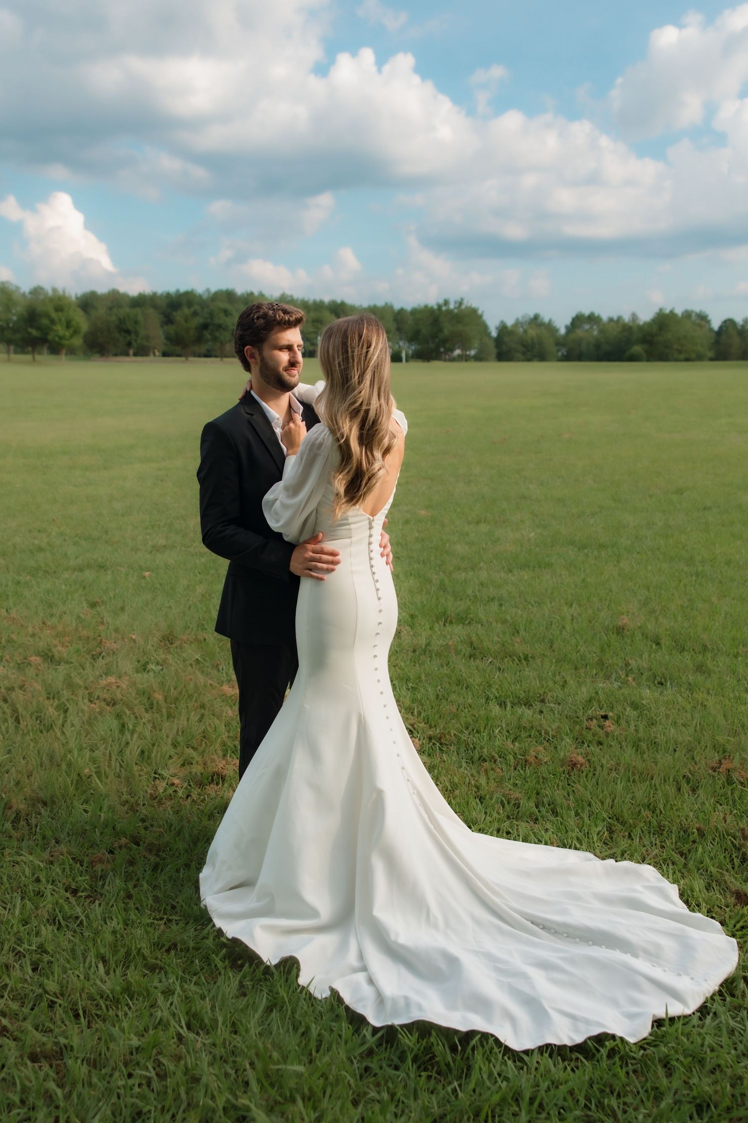Bride in elegant white mermaid gown with button back train poses with groom in black suit in green field.