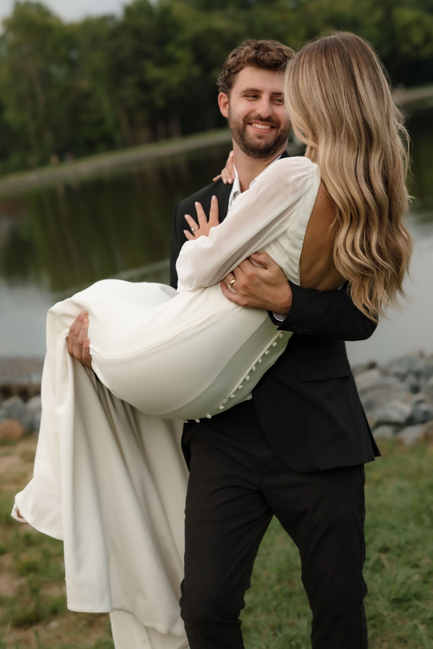 Smiling groom in black suit lifts bride in white backless gown near a lakeside setting.