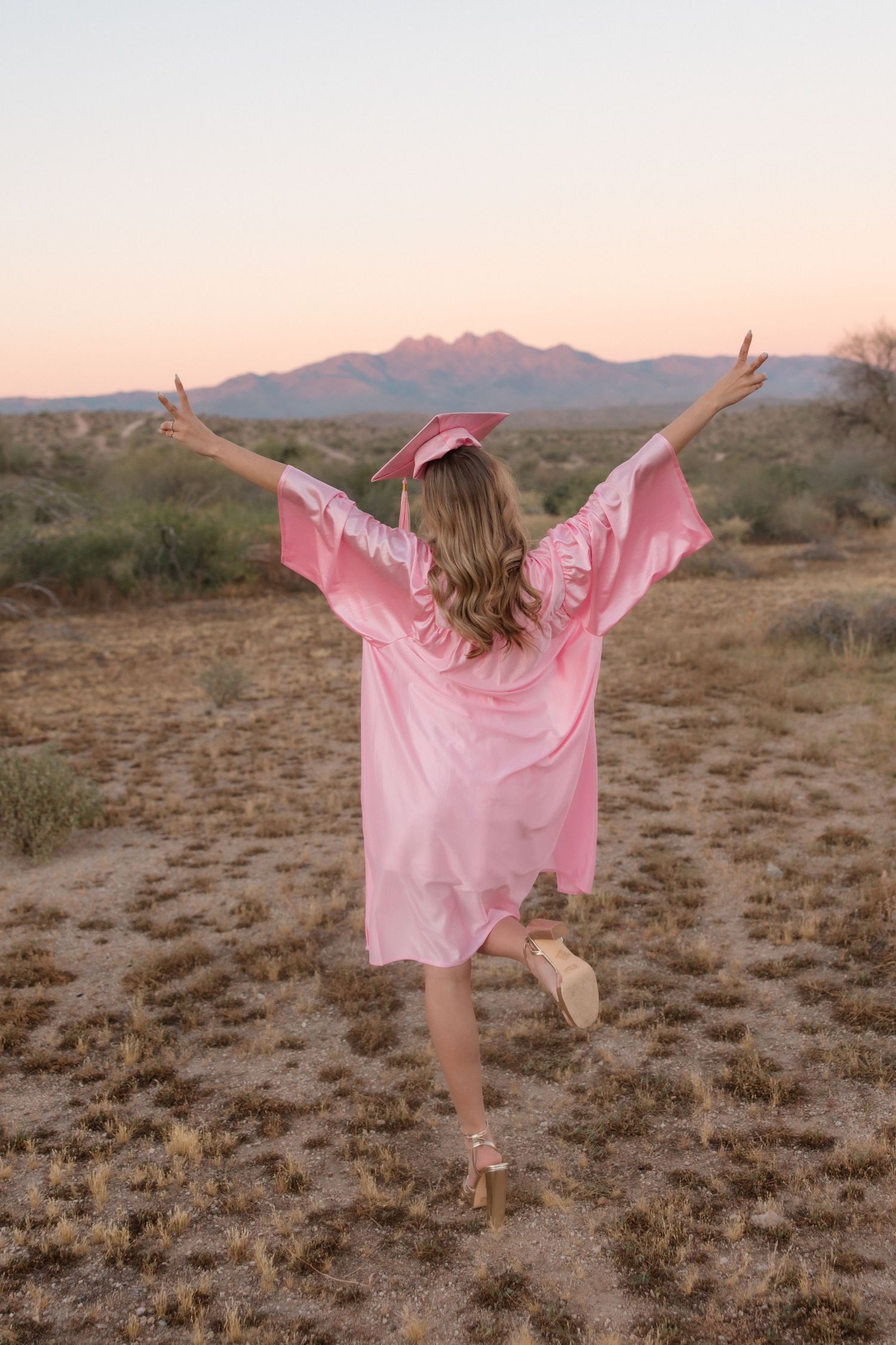 Woman in pink graduation cap and gown celebrates outdoors with arms raised, desert mountains at sunset behind her.