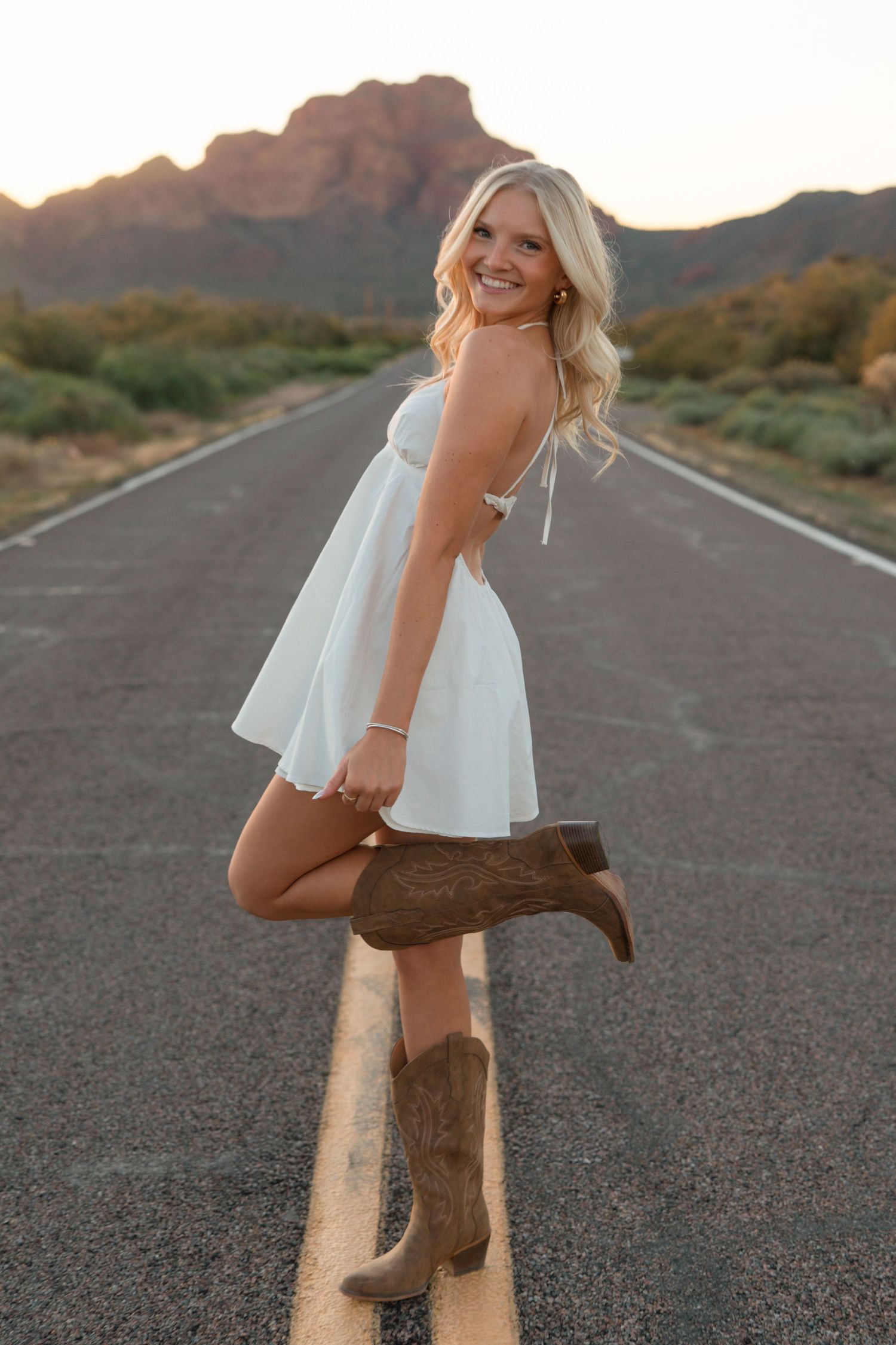 Blonde woman in white dress and cowboy boots posing on desert road at sunset with mountain backdrop.