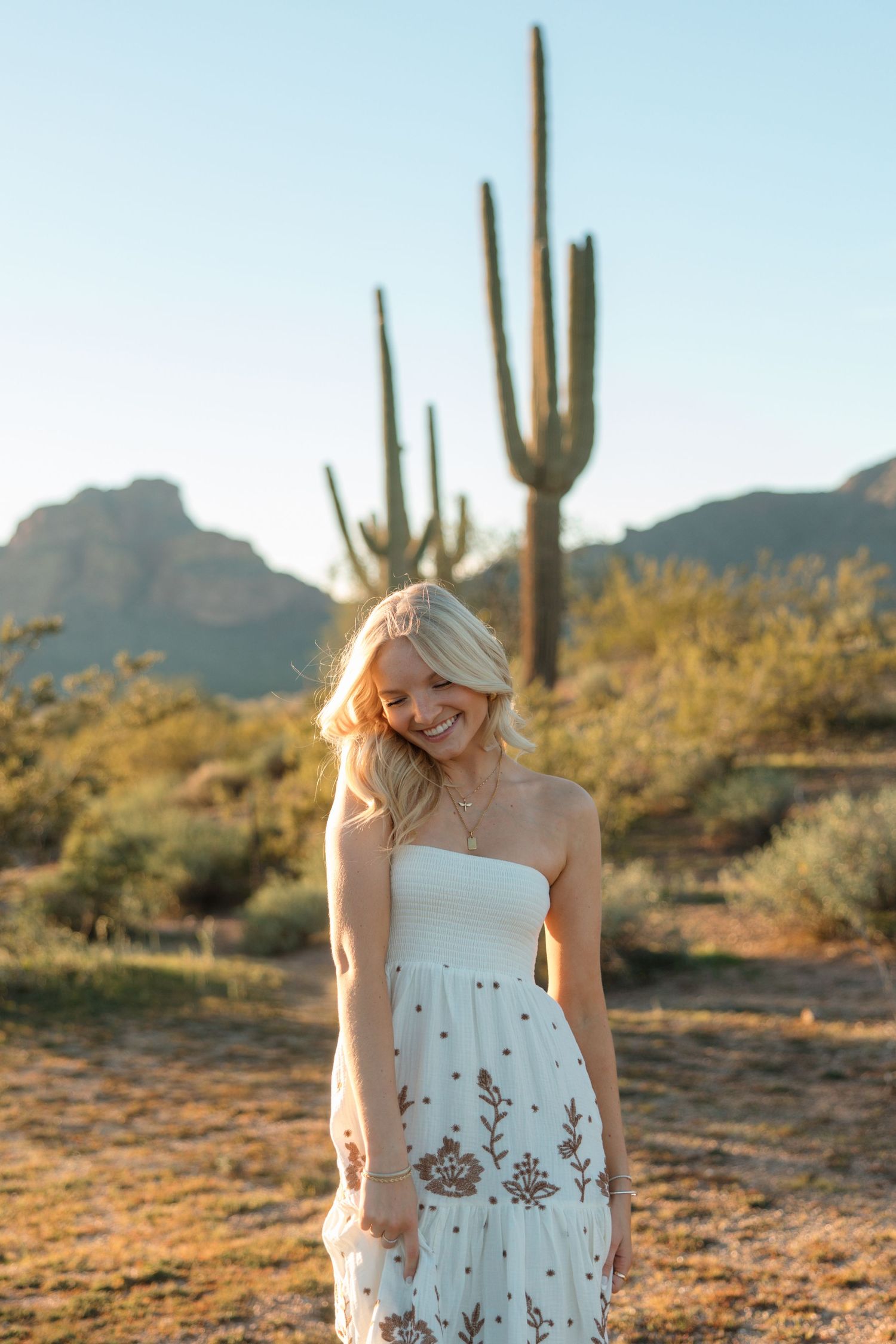 Smiling blonde woman in white strapless dress standing in Arizona desert with saguaro cacti and mountains.