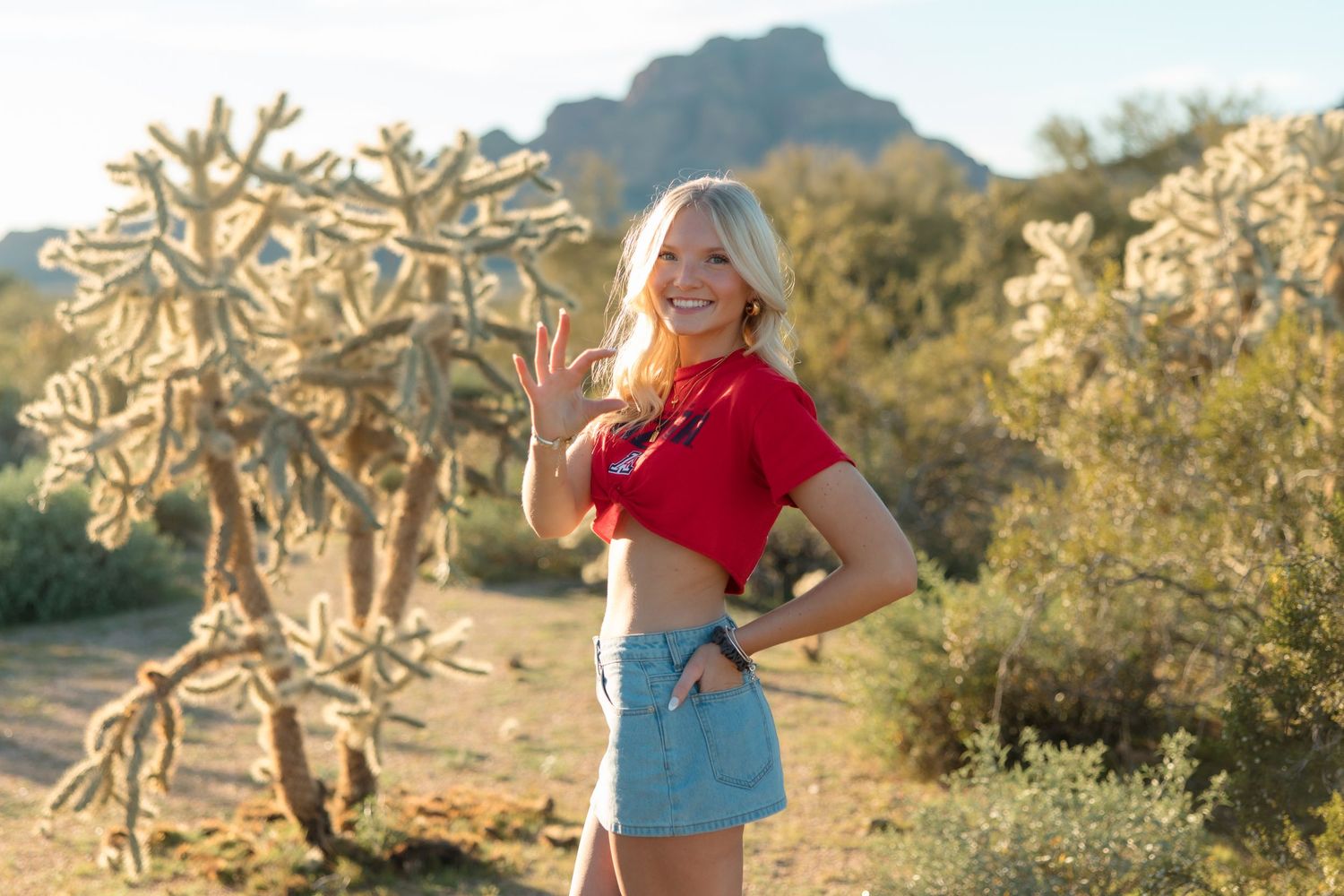 Blonde woman in red crop top and denim skirt waving near cholla cactus in sunny desert landscape.