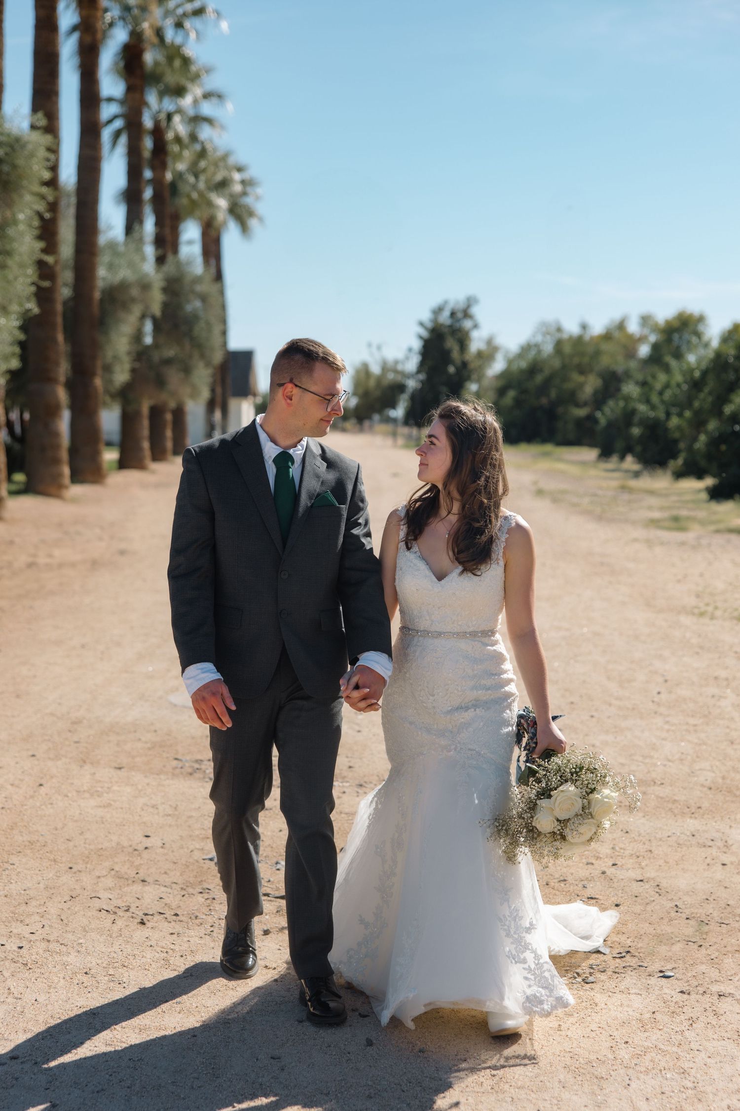Bride and groom walk hand-in-hand along a palm tree-lined path on their wedding day.