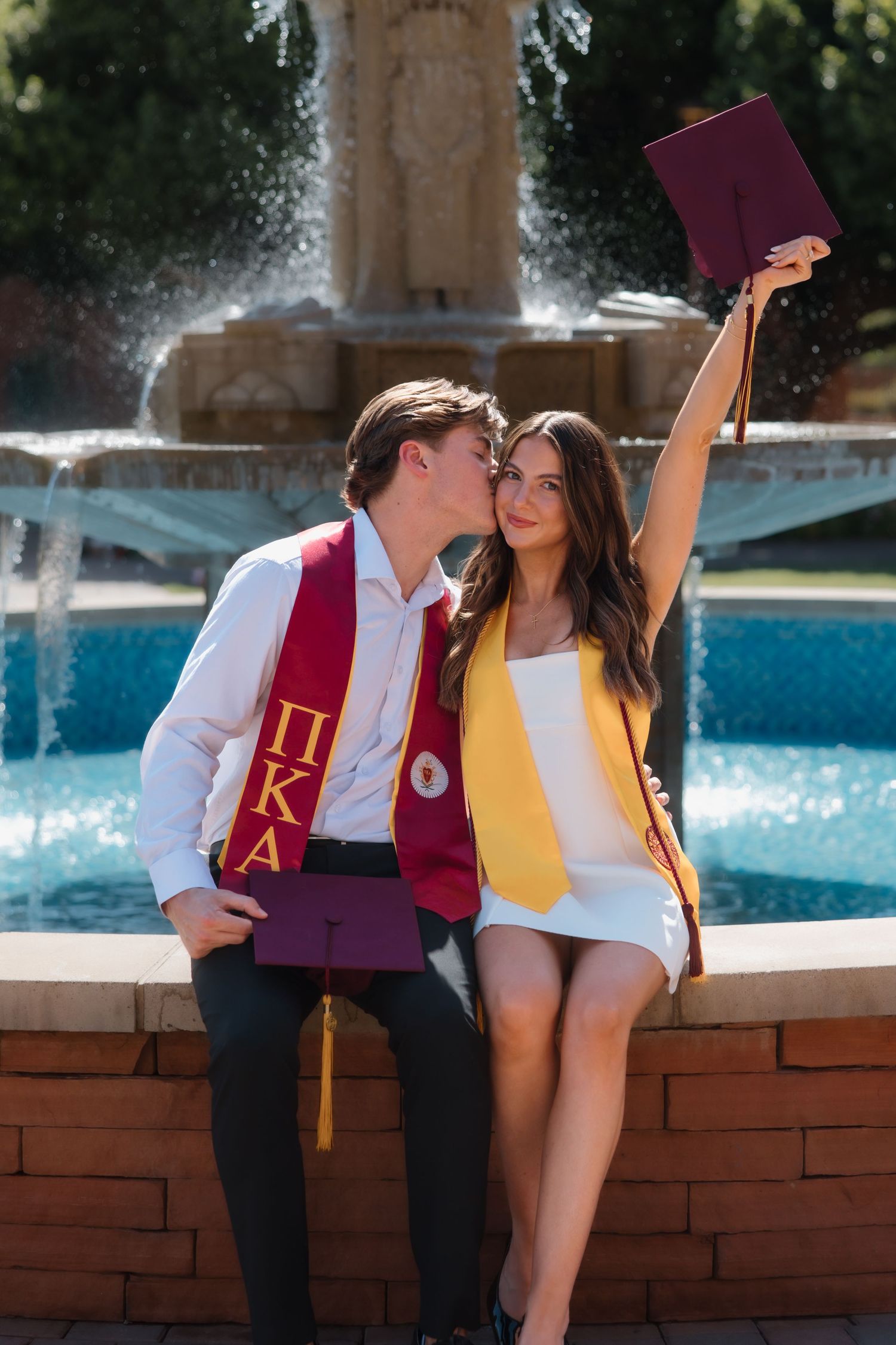 Graduate kisses partner on cheek as she raises cap triumphantly by campus fountain celebration.