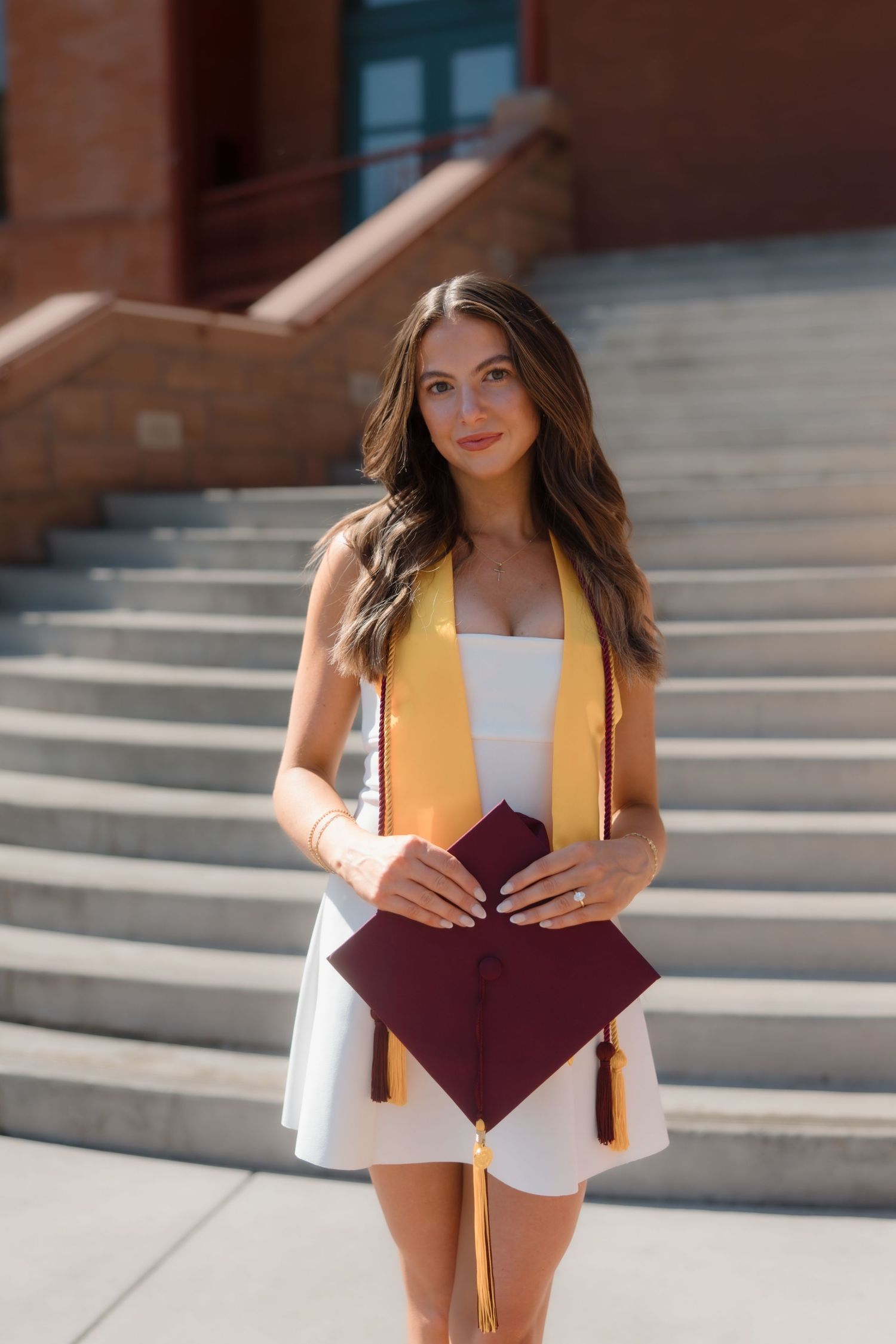 Young woman in white dress holding maroon graduation cap with gold ASU stole at Arizona State University.