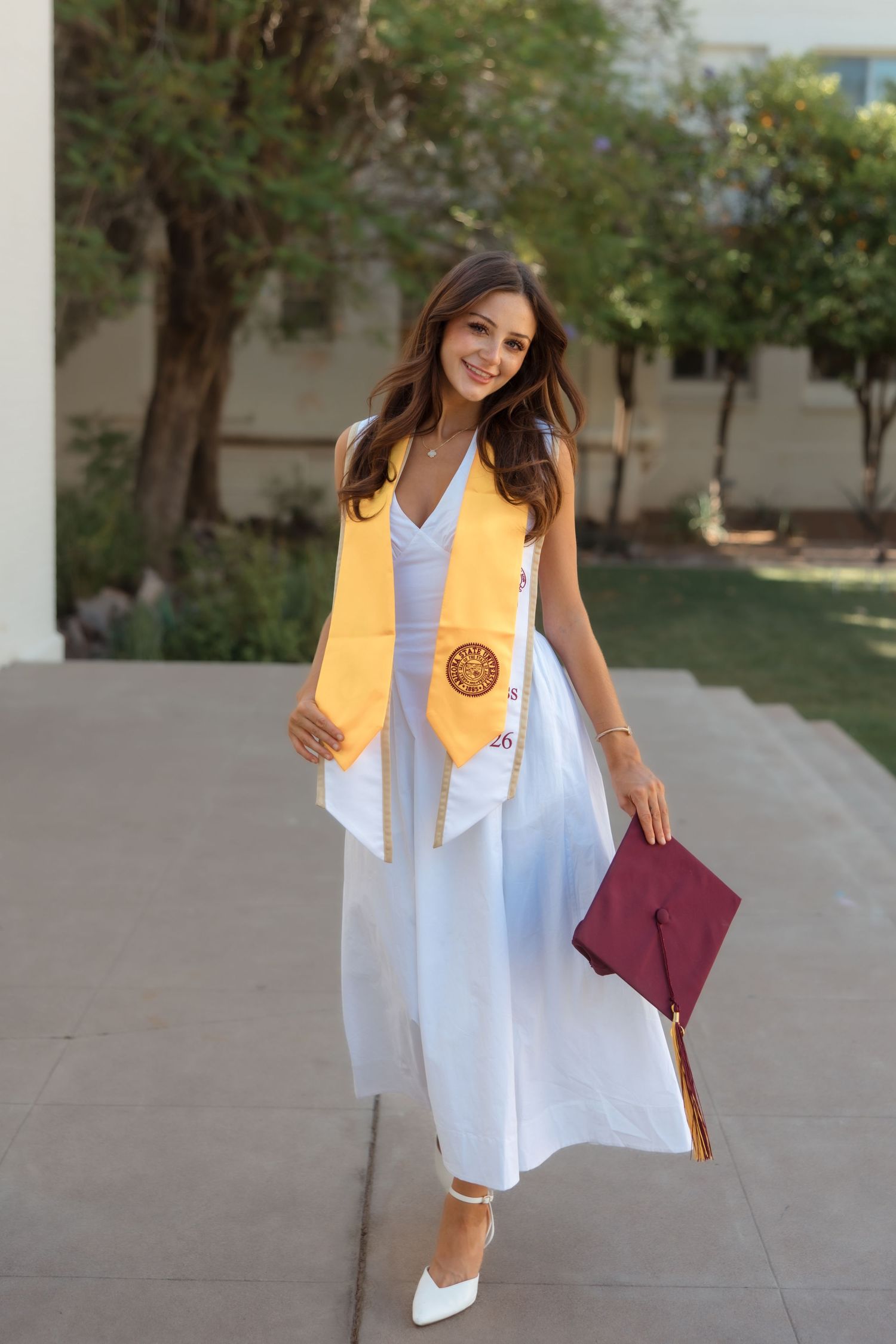 Smiling graduate in white dress with gold honor stole holds maroon cap on sunny campus walkway.