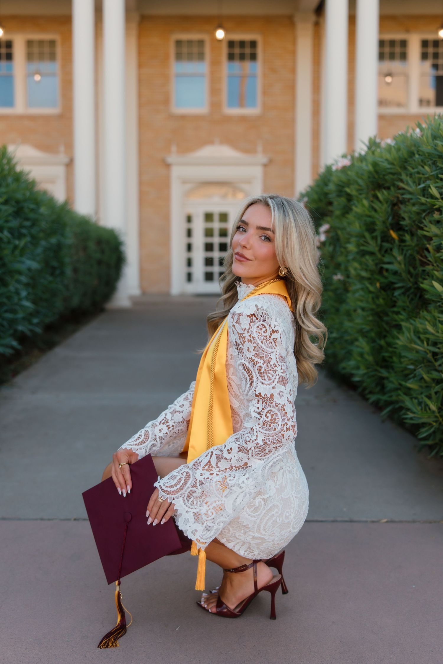 Blonde woman in white lace dress with gold sash holds maroon graduation cap outside brick building.