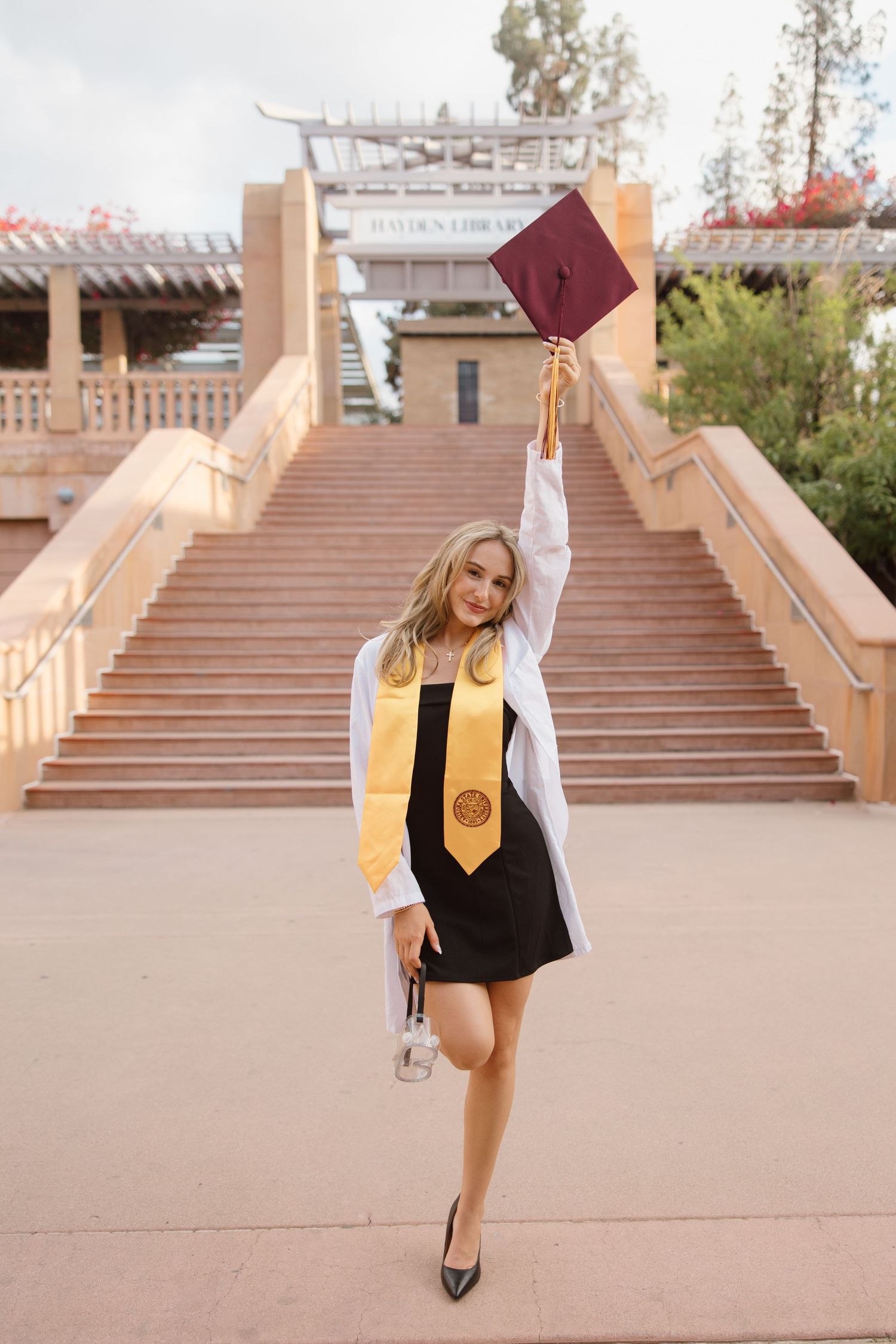 Young woman in white graduation gown and gold stole raises maroon cap on university campus steps.