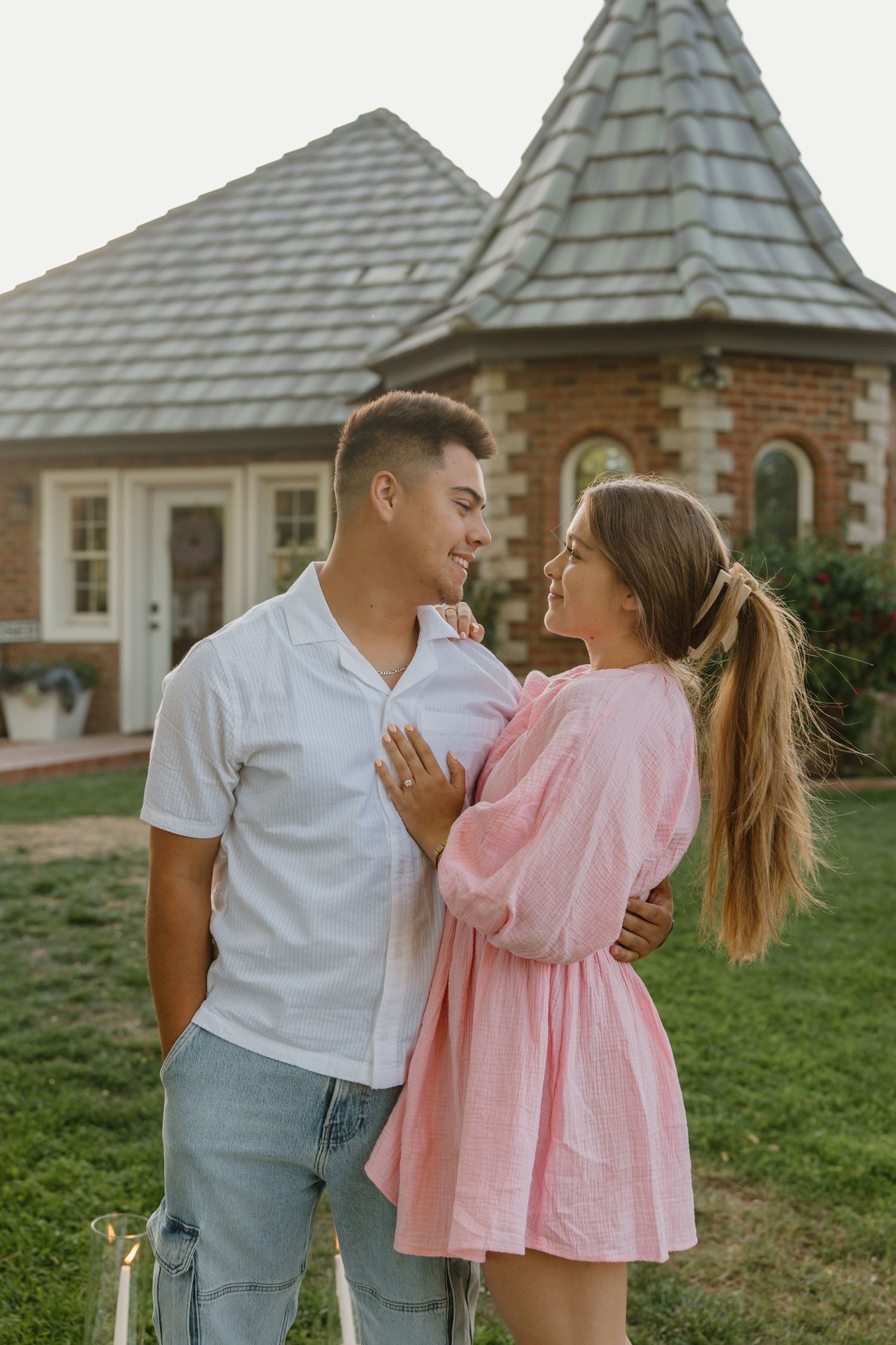 A young couple in casual attire embracing and smiling at each other in front of a charming brick cottage.