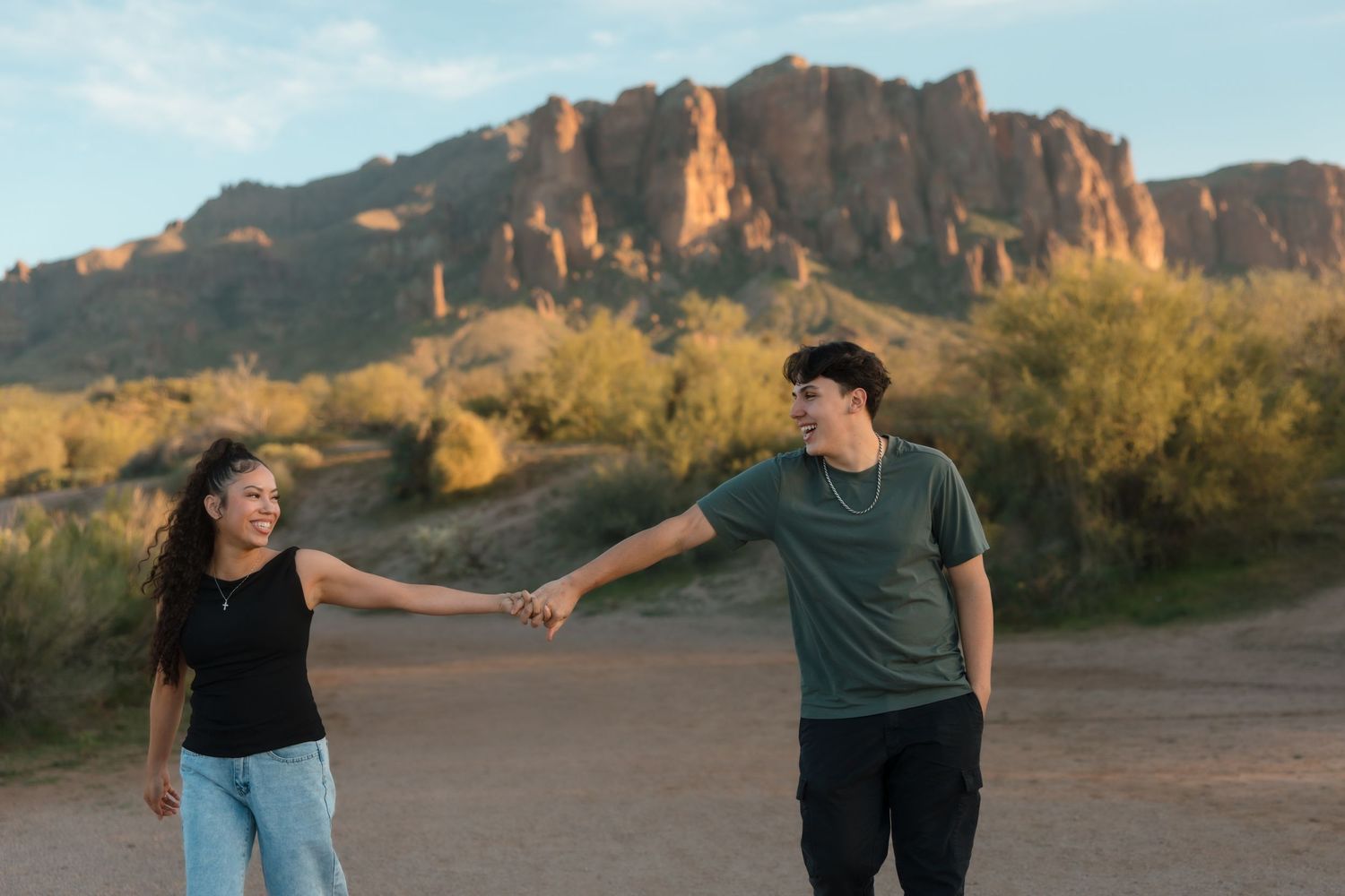 A happy couple holds hands and laughs together in a desert setting with rugged mountains behind them.
