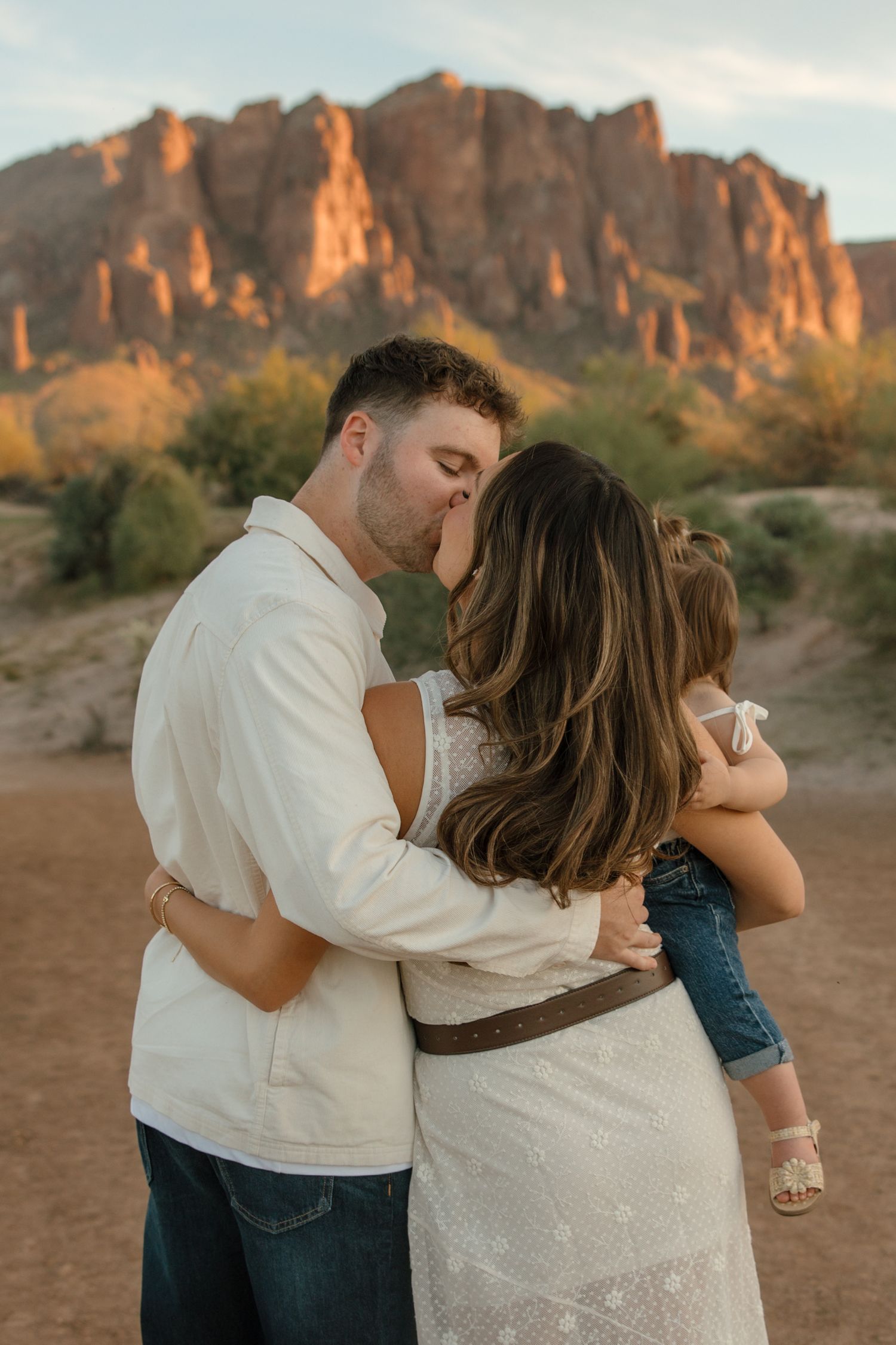 Couple sharing a romantic kiss while holding their young child in front of Arizona desert mountains.