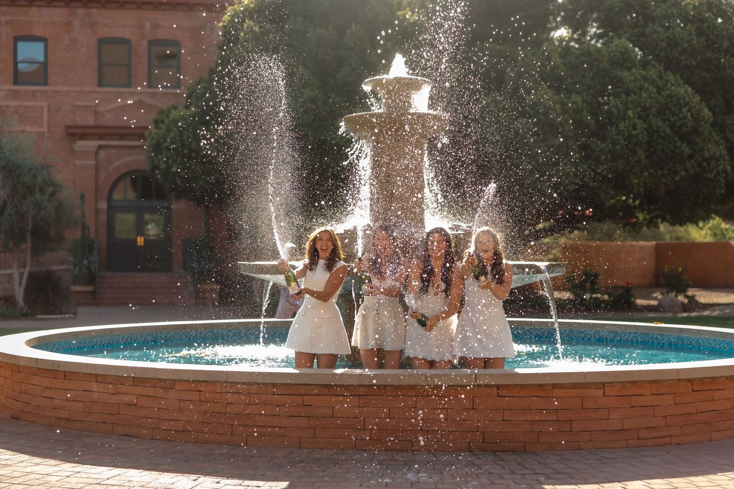 Three women in white dresses celebrate with confetti inside a large outdoor campus fountain.