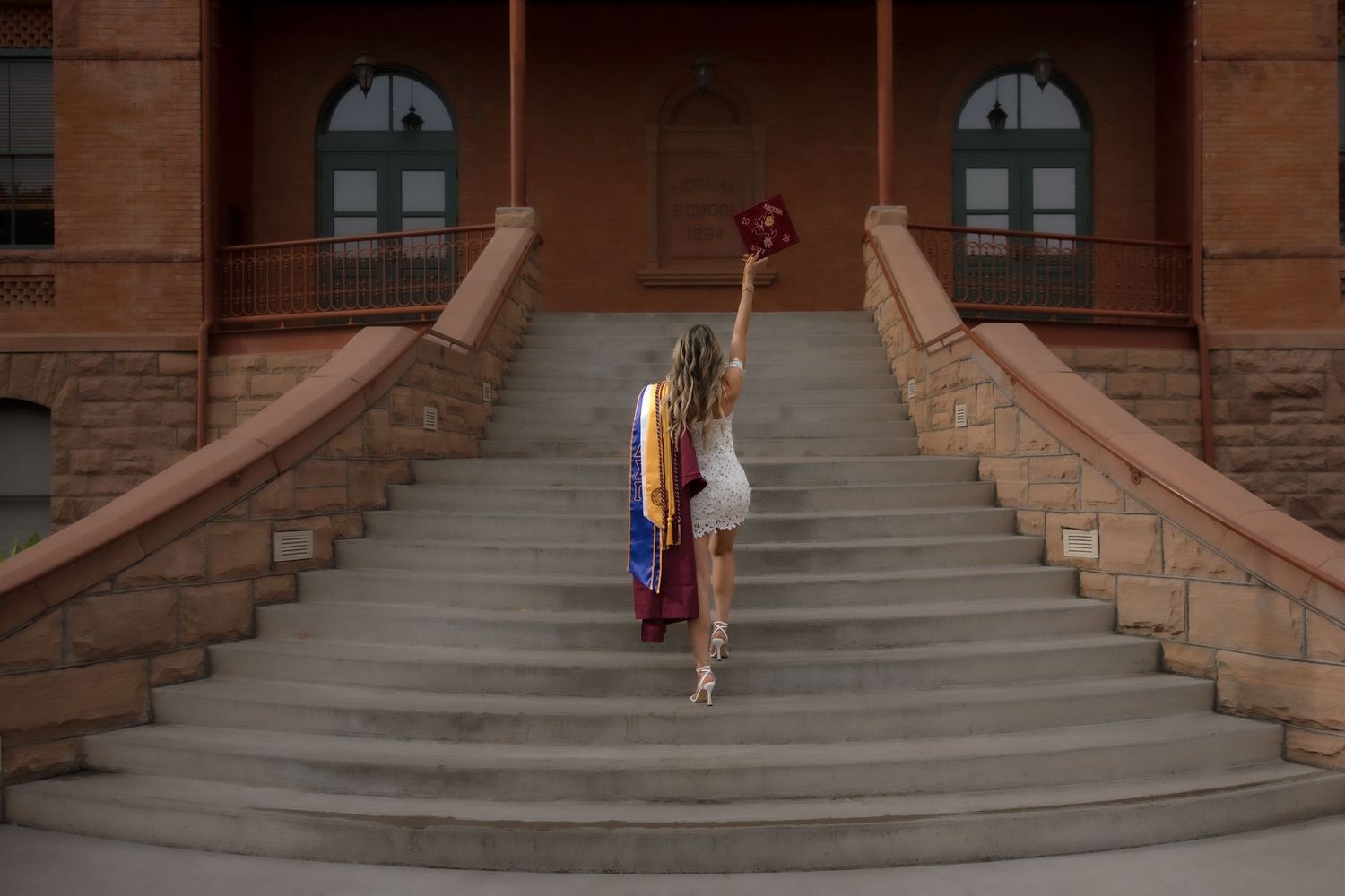 ASU graduate walking up grand staircase of historic Old Main building holding maroon graduation cap and colorful sashes.