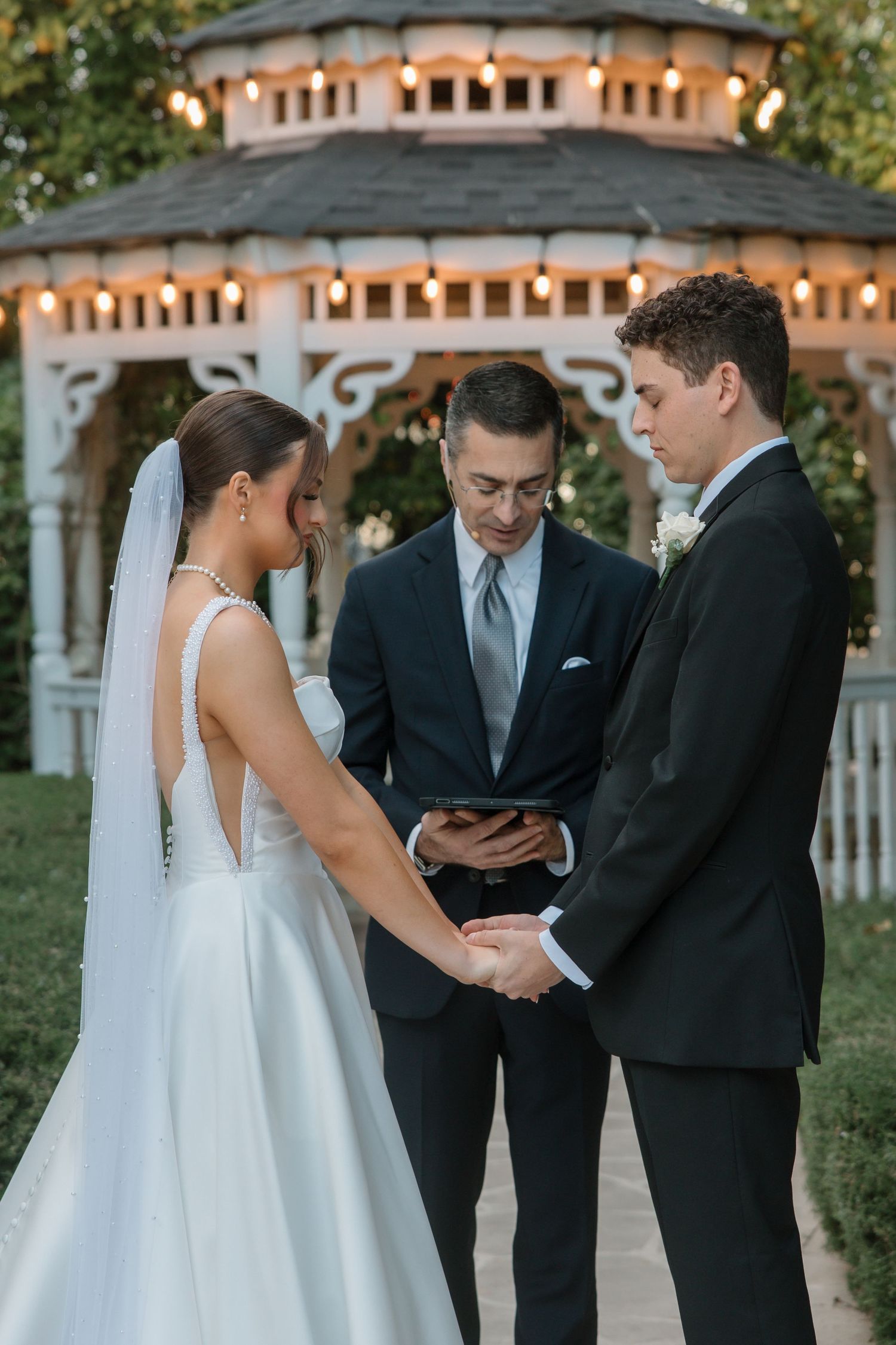 Bride and groom hold hands during outdoor wedding ceremony at a gazebo while officiant reads vows.