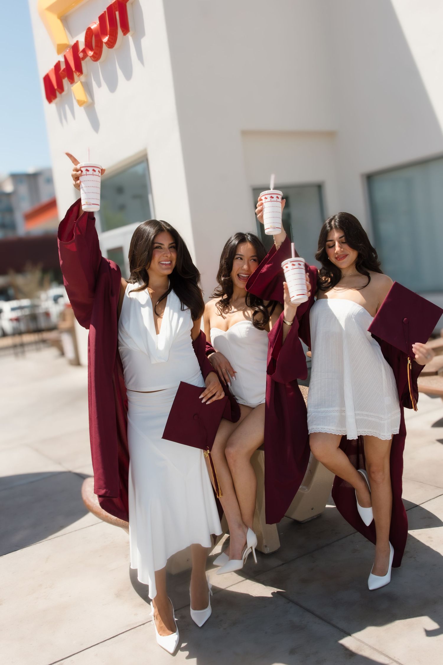 Three excited female graduates in burgundy caps and gowns raise In-N-Out drinks outside the restaurant.