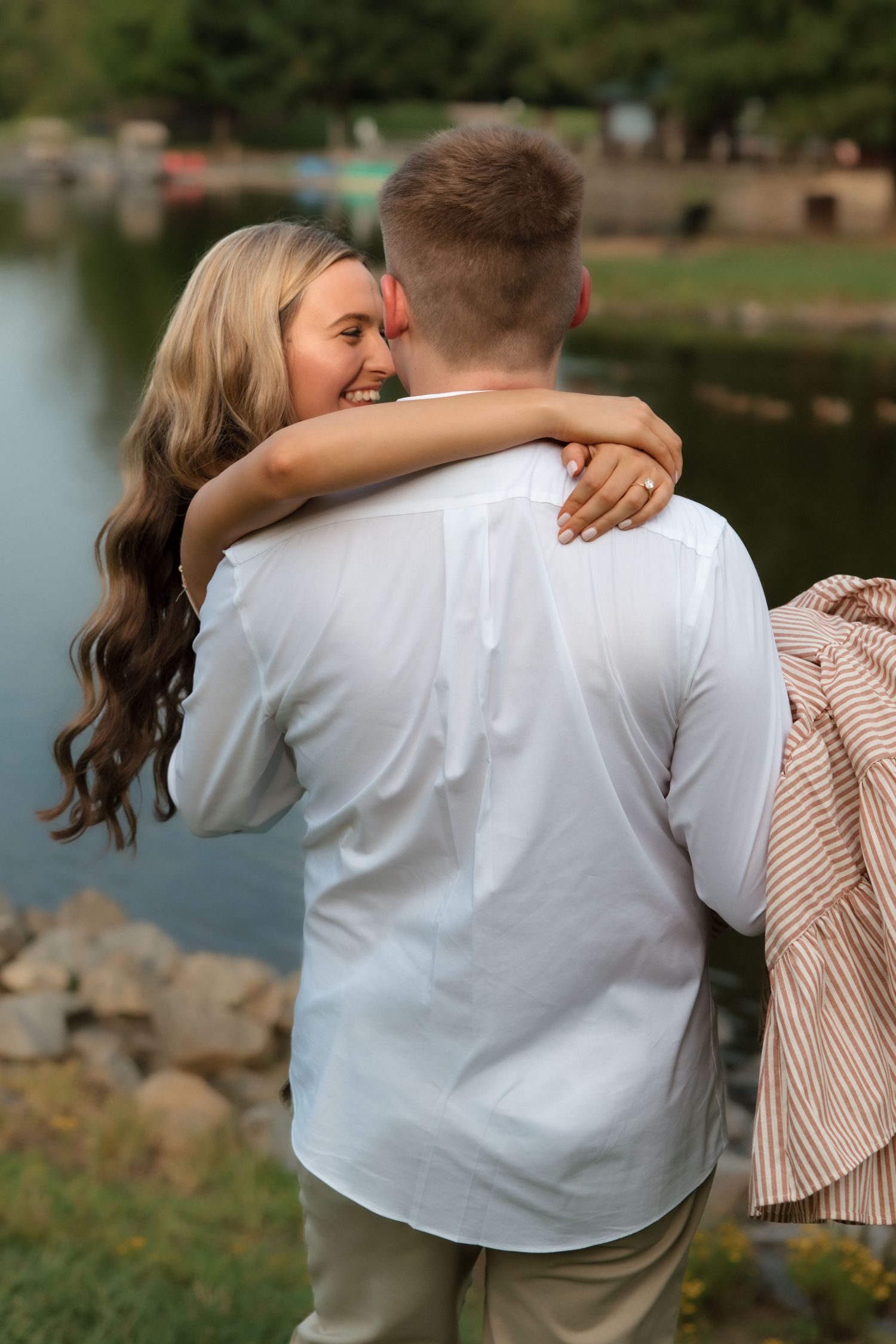 A couple embraces and laughs near a serene lake, the woman wearing a ring as they share a romantic outdoor moment.