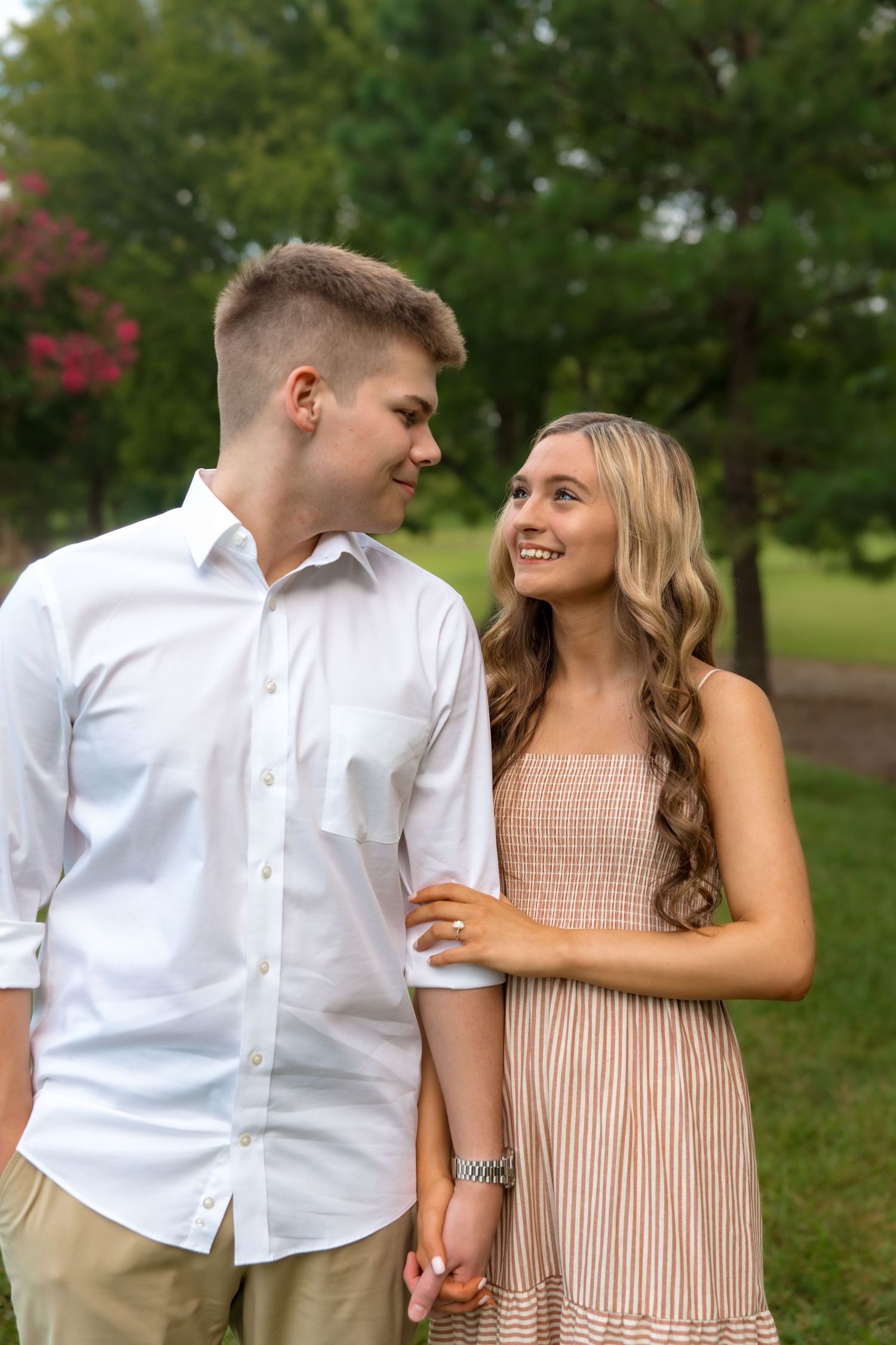 Young couple holding hands and smiling at each other outdoors in a lush green park setting.