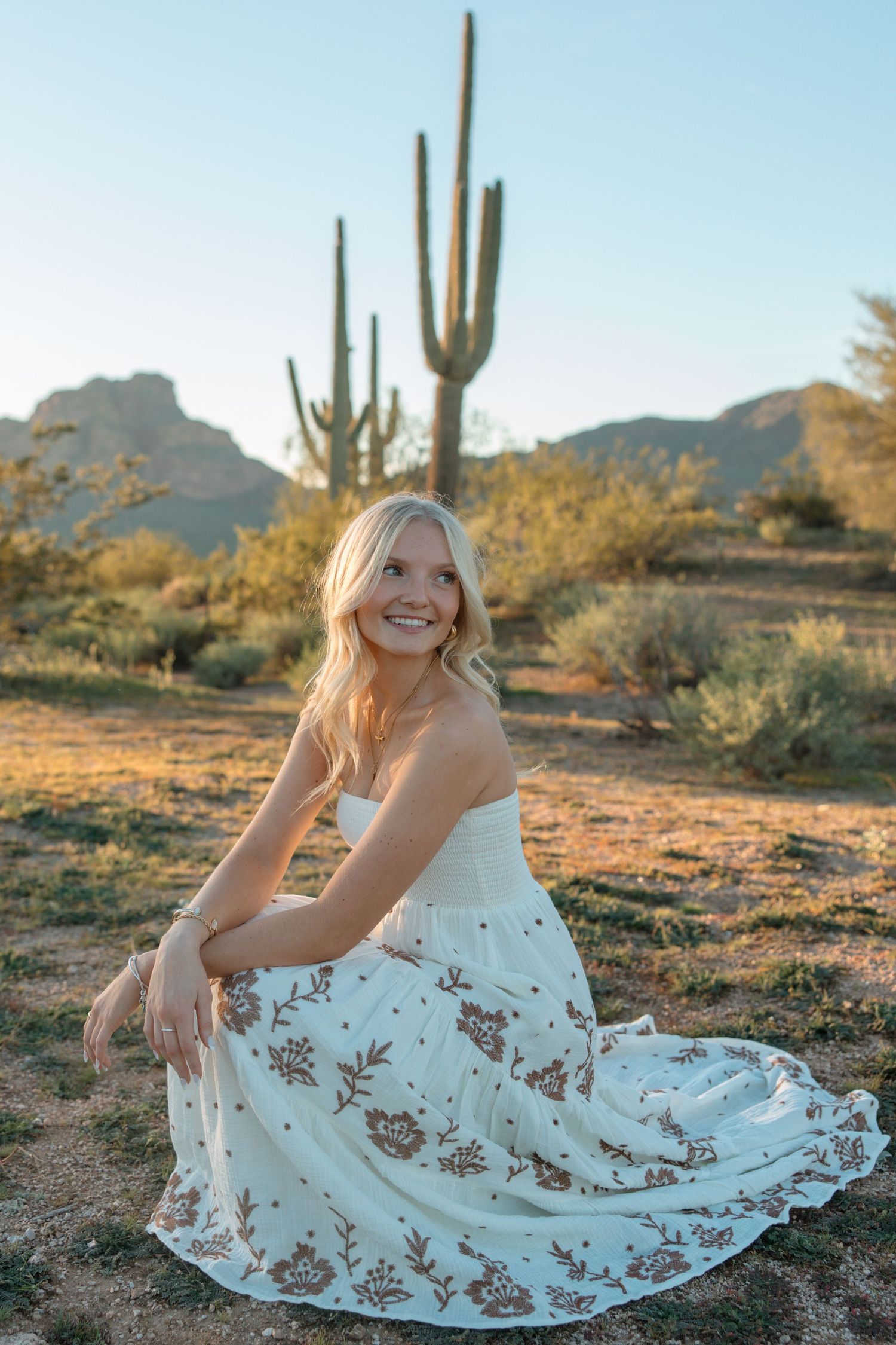 Smiling blonde woman in floral white dress sitting in Arizona desert with saguaro cactus and mountains at sunset.