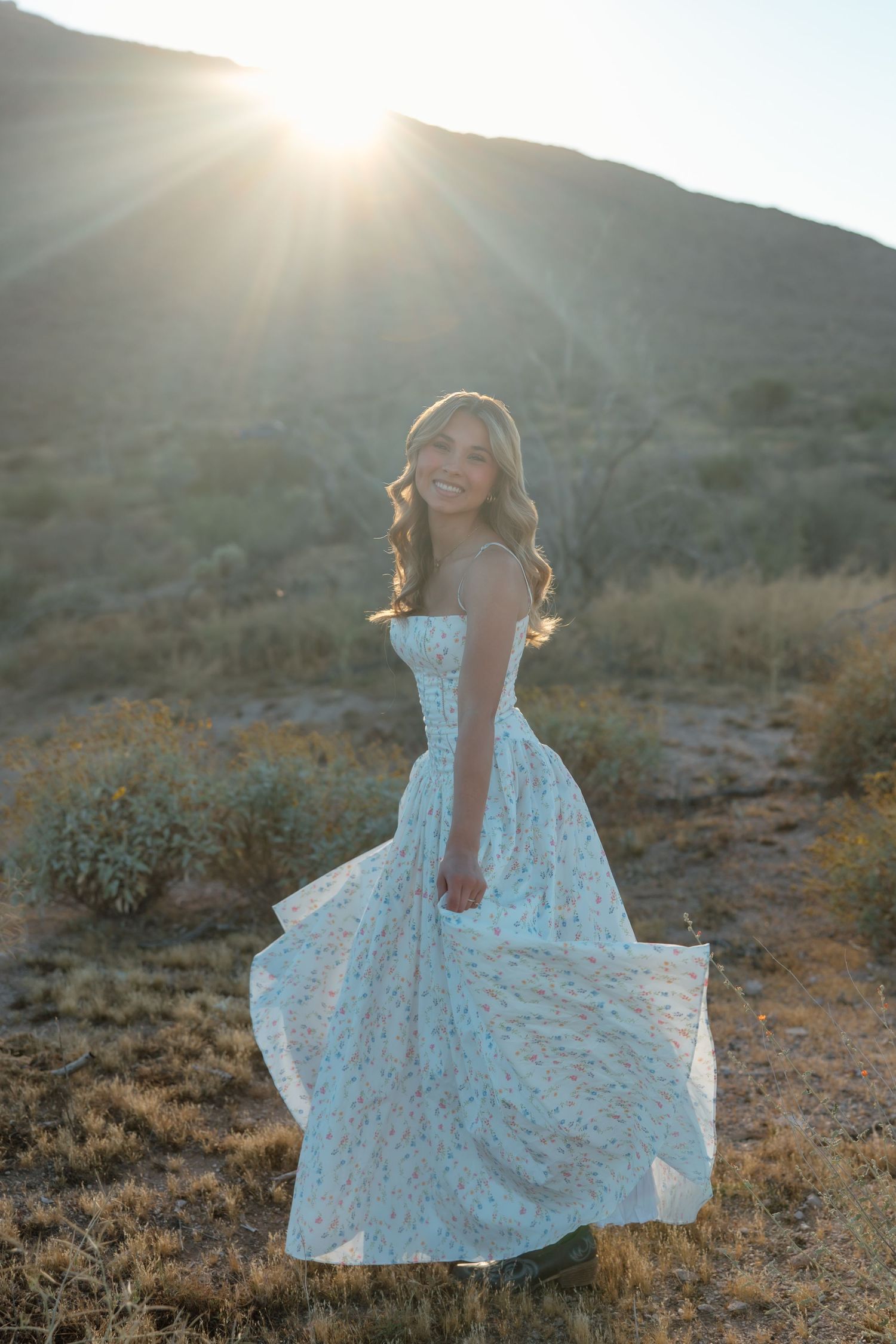 Young woman in floral gown twirling at golden hour with sun rays over desert hills.