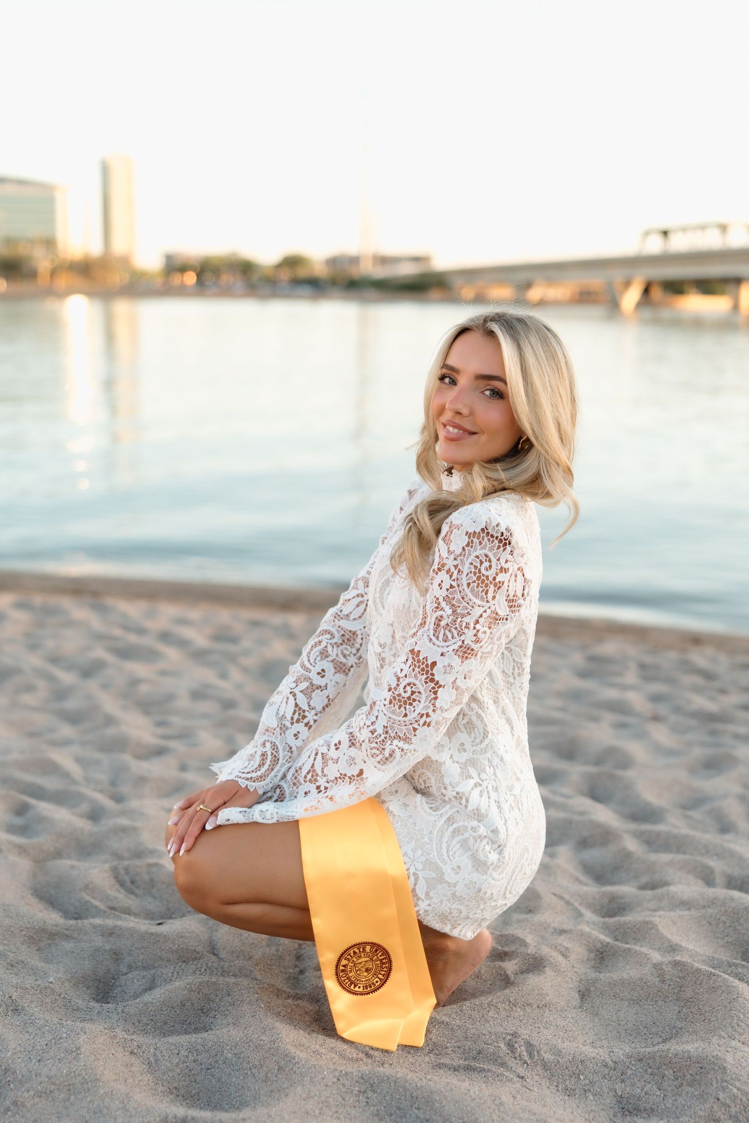 Blonde woman in white lace dress sits barefoot on sandy beach with gold graduation stole.