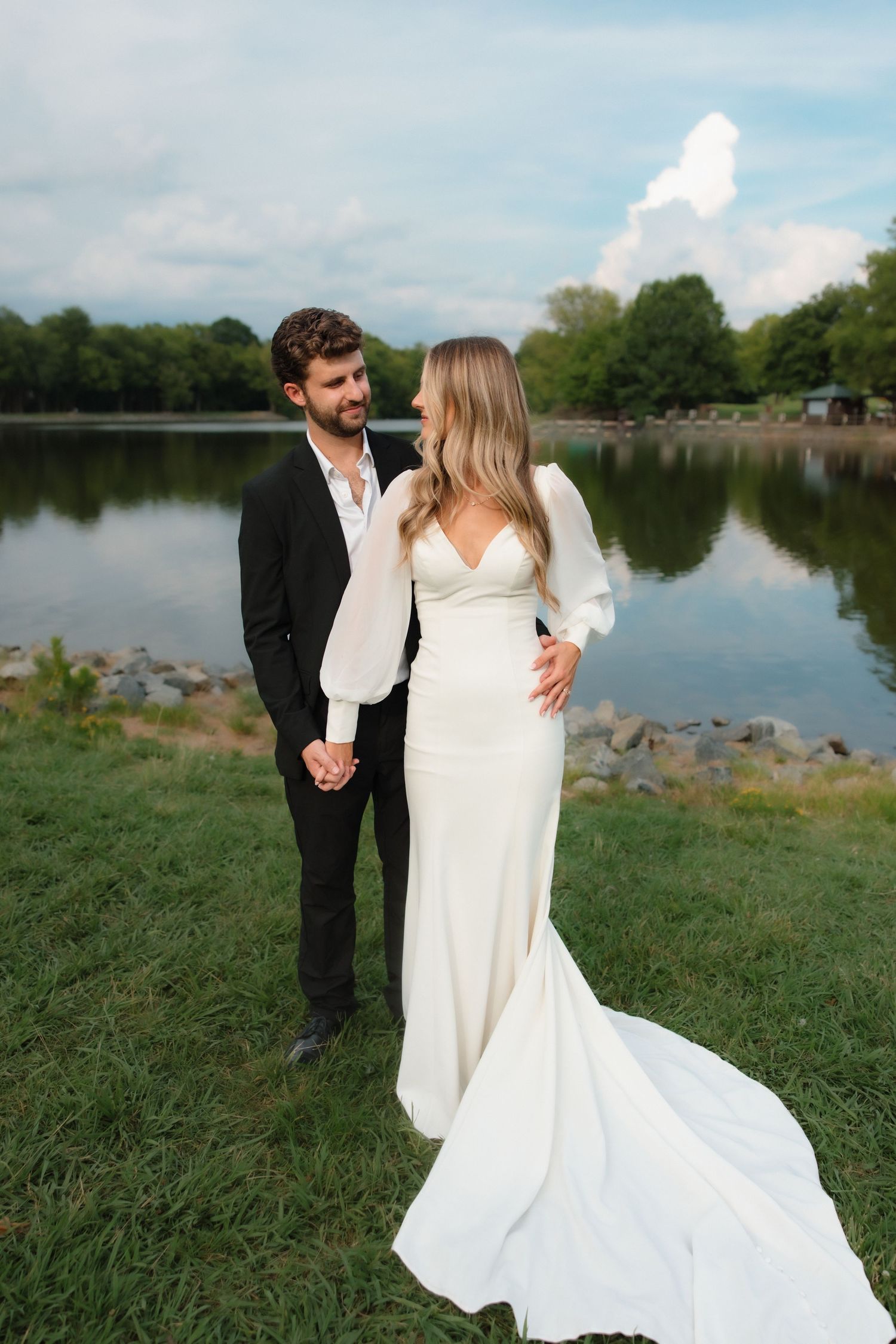 Wedding couple holding hands by a serene lake, bride in elegant white gown with long train.