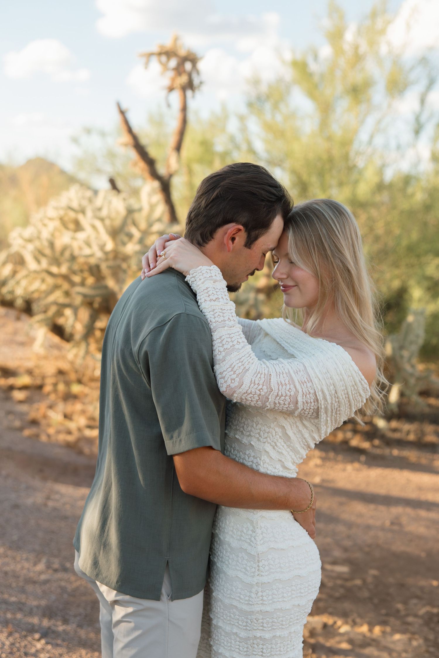 Couple embracing in a desert landscape with cacti, woman in white knit dress and man in sage green shirt.